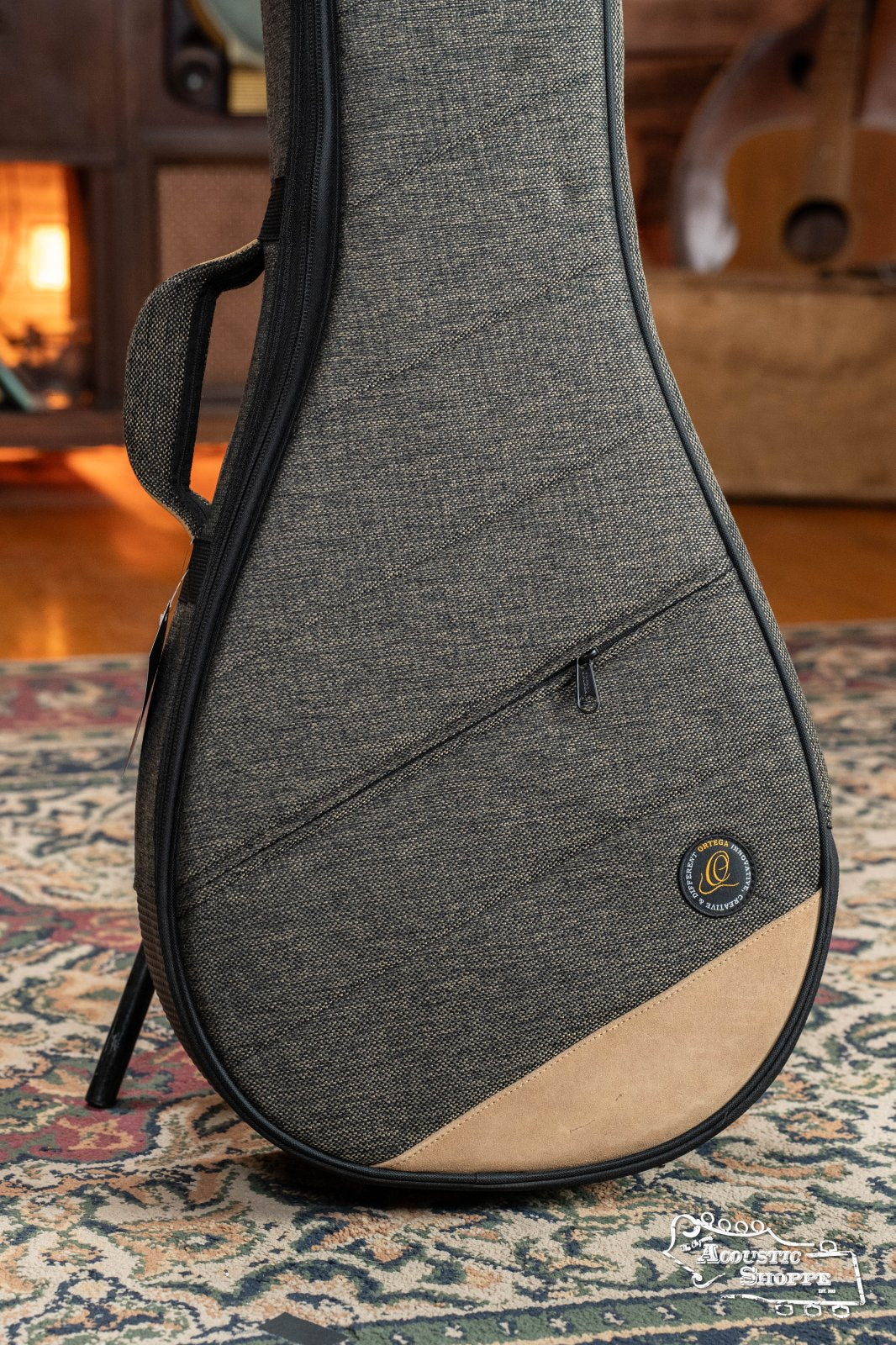 The Ortega OSOCABJ-MO Banjo Soft Case in Mocca by Ortega, featuring gray fabric, a zipper pocket, and tan accents, stands on a patterned rug with a guitar and lit fireplace partially visible in the background.