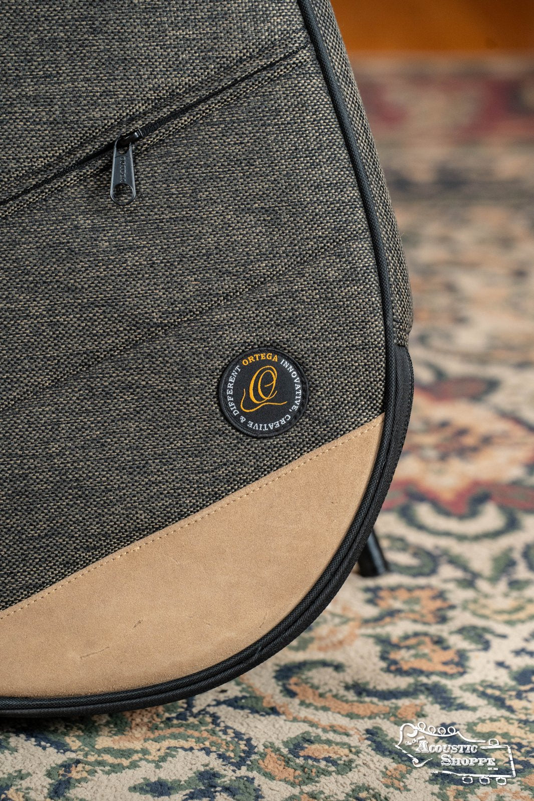 Close-up of the Ortega OSOCABJ-MO Banjo Soft Case in Mocca with tan and gray padded fabric, a round Ortega logo patch, black zipper, and textured finish, displayed on a patterned carpet.