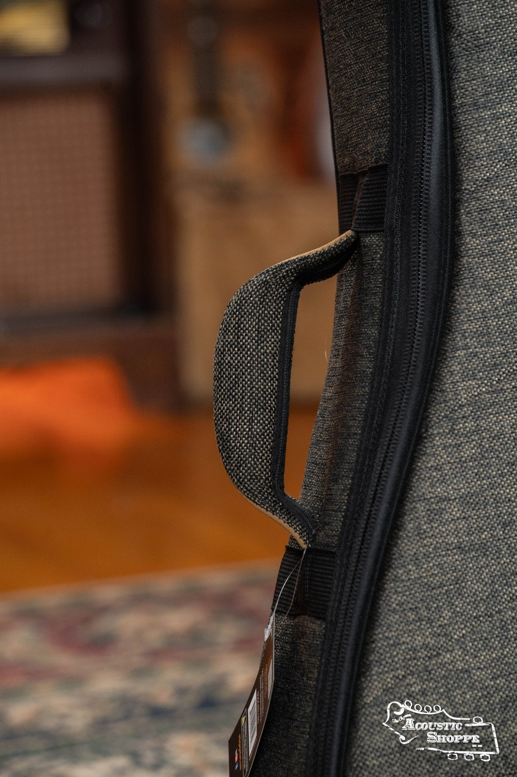Close-up of the padded handle on the Ortega OSOCABJ-MO Banjo Soft Case in Mocca, with a blurred indoor backdrop featuring a patterned rug and wood floor. The Ortega logo is visible in the lower right corner.