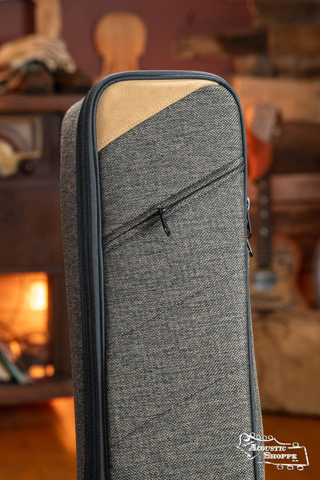 The Ortega OSOCABJ-MO Banjo Soft Case in Mocca, a padded gig bag, stands upright in a cozy room beside a gray fabric guitar case with tan accents, with blurred musical instruments and a fireplace in the background.