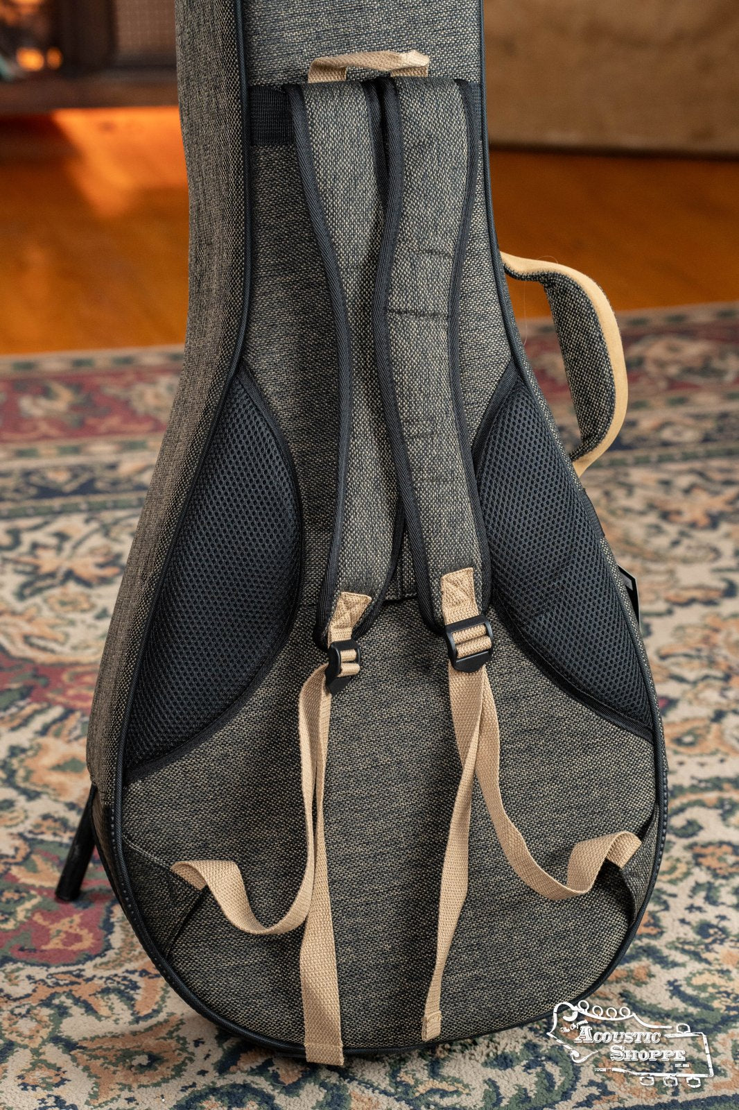 The Ortega OSOCABJ-MO Banjo Soft Case in Mocca, by Ortega, stands upright on a patterned rug. It features beige and black fabric, reinforced black mesh areas, padding, and backpack-style straps.