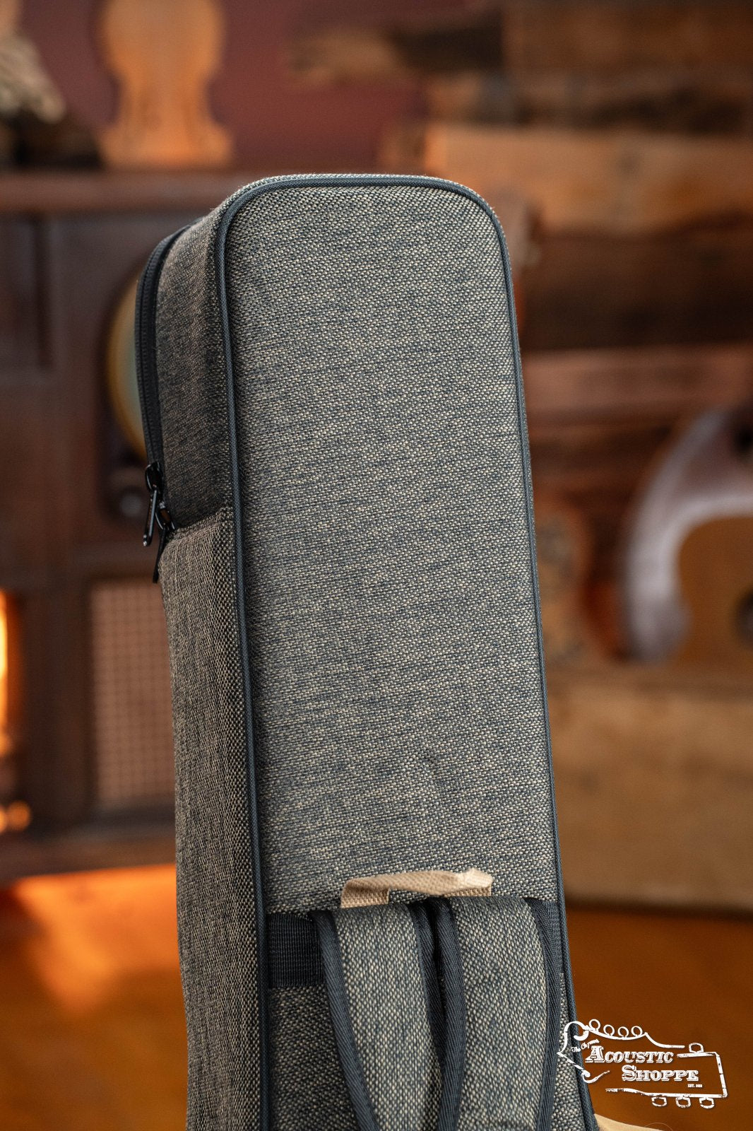 A close-up of the Ortega OSOCABJ-MO Banjo Soft Case in Mocca, featuring padded textured fabric and a zipper, stands upright indoors with blurred wooden furniture and instruments in the background. Ortega branding is visible in the bottom right corner.