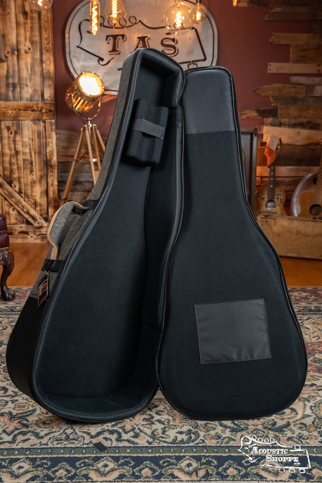 The Ortega OSOCADN-MO Dreadnought Guitar Soft Case in Mocca stands upright on a patterned rug in a cozy, rustic room with wooden walls, warm lighting, and musical instruments in the background.
