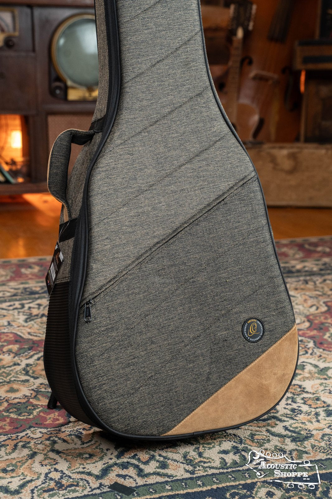 The Ortega OSOCADN-MO Dreadnought Guitar Soft Case in Mocca stands upright on a patterned rug in a cozy, warmly lit room featuring vintage décor. Its brown accents and padded design highlight Ortega’s signature style.