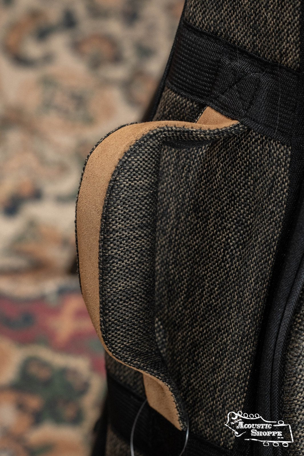 Close-up of the Ortega OSOCADN-MO Dreadnought Guitar Soft Case in Mocca, showing its brown and black padded design with tan accents, side handle, black straps, and the Acoustic Shoppe logo on a multicolored carpet background.
