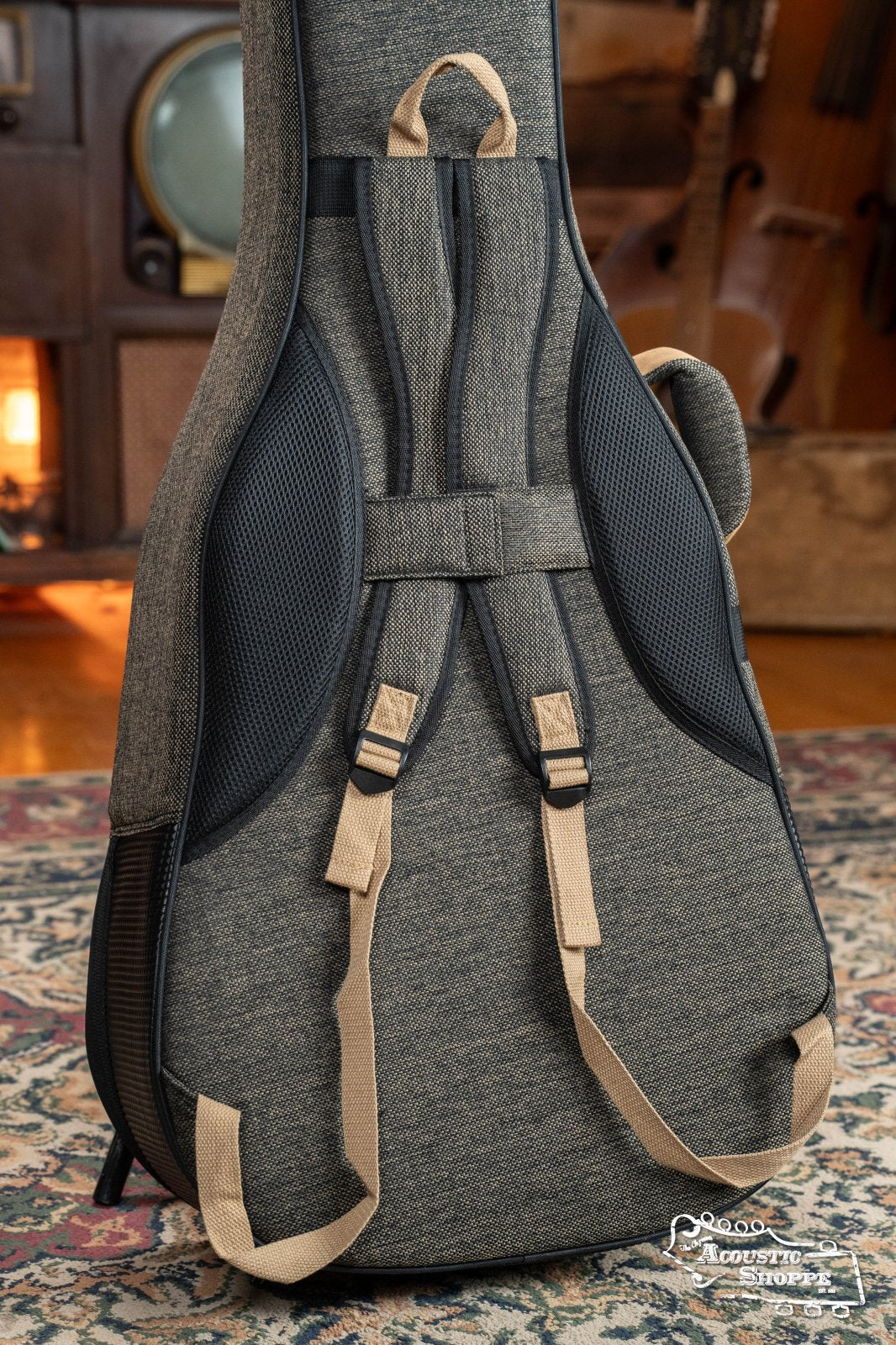 The Ortega OSOCADN-MO Dreadnought Guitar Soft Case in Mocca, with textured gray material, tan straps, and black accents, stands upright on a patterned rug; vintage furniture is softly blurred in the background.