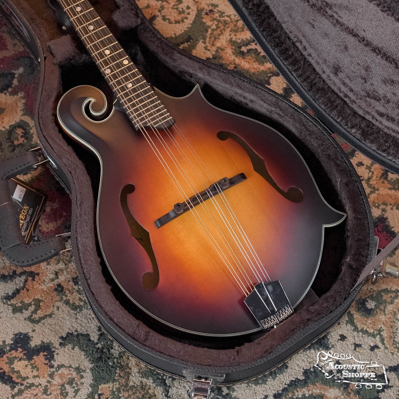 An F-style sunburst mandolin with scroll and f-holes lies in an open Ortega OMCSTD-F Hardshell Mandolin Case by Ortega, showing its metal tailpiece and a case tag, set on a patterned carpet.
