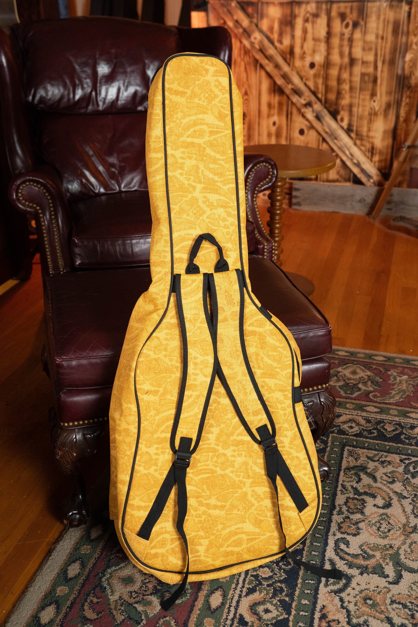 An Ortega OGBAC-DN-SUJ Dreadnought Guitar Gigbag in Sun Jean rests upright on a brown leather armchair in a cozy room with wooden floors, a patterned rug, and wood-paneled walls.