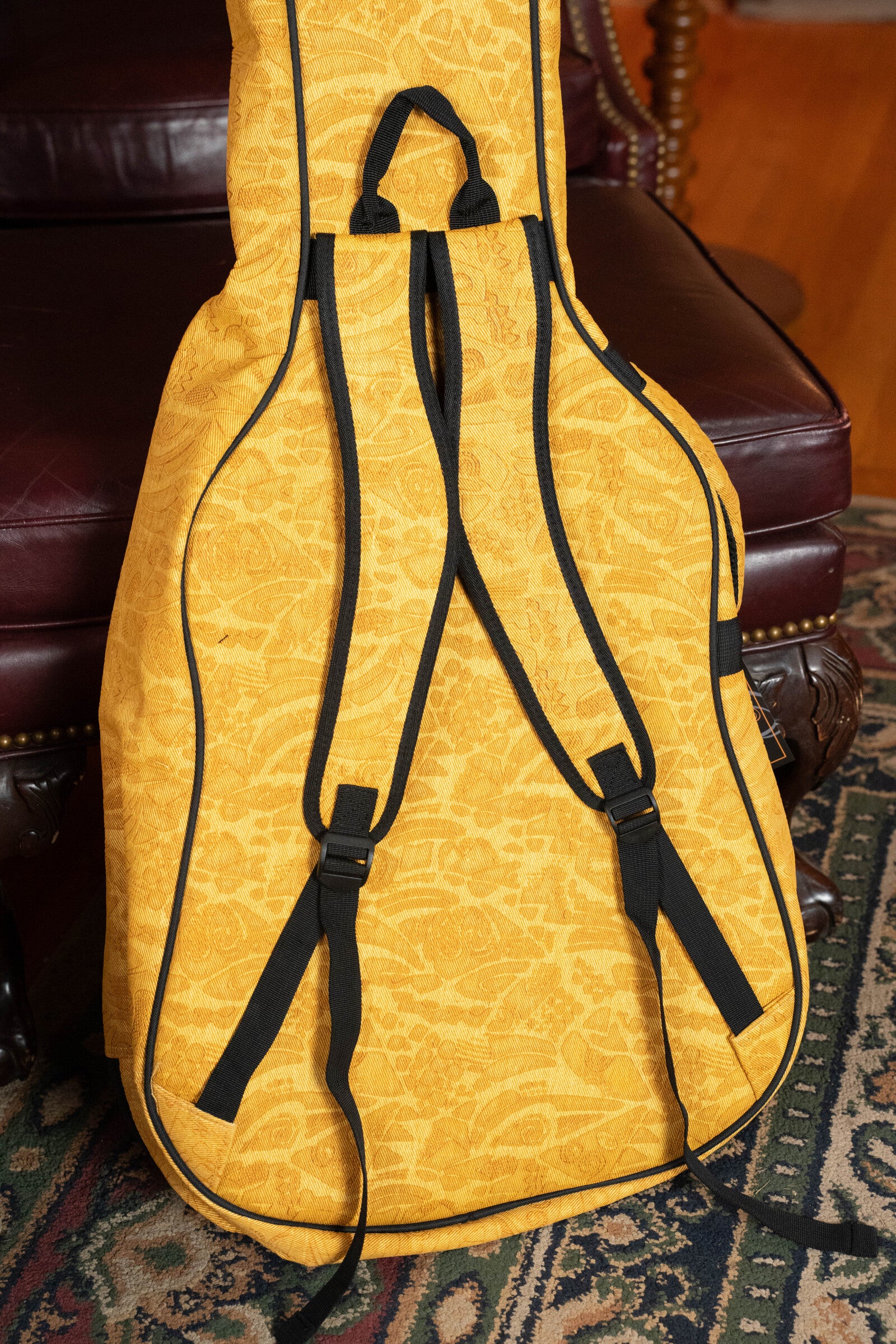 The Ortega OGBAC-DN-SUJ Dreadnought Guitar Gigbag - Sun Jean, a yellow patterned case with black straps, stands upright on a patterned rug, leaning against a dark leather chair with wooden legs.