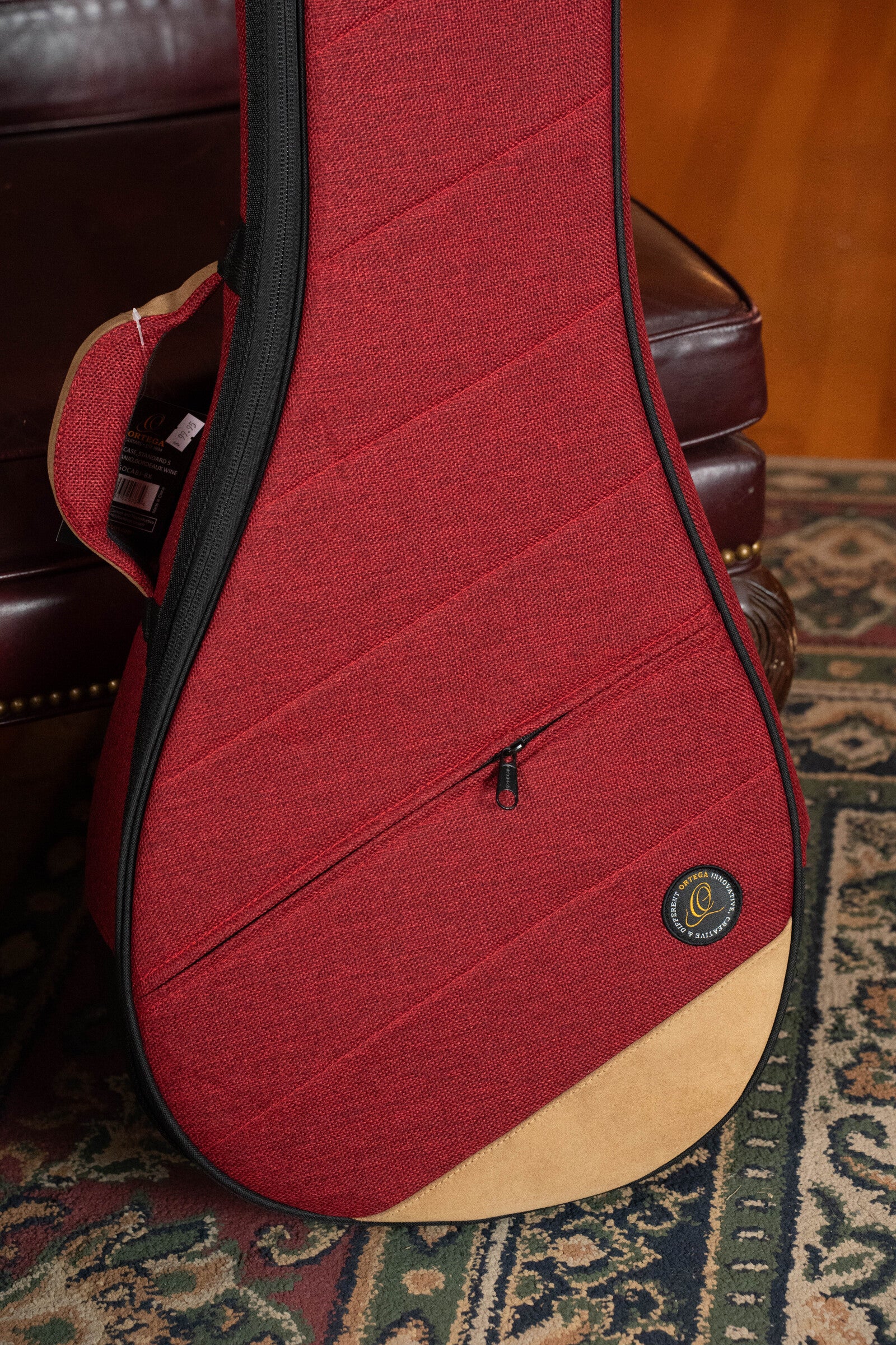 The Ortega OSOCABJ-BX Banjo Soft Case in Bordeaux Wine leans against a brown leather chair, partially resting on a patterned rug with green and red tones.