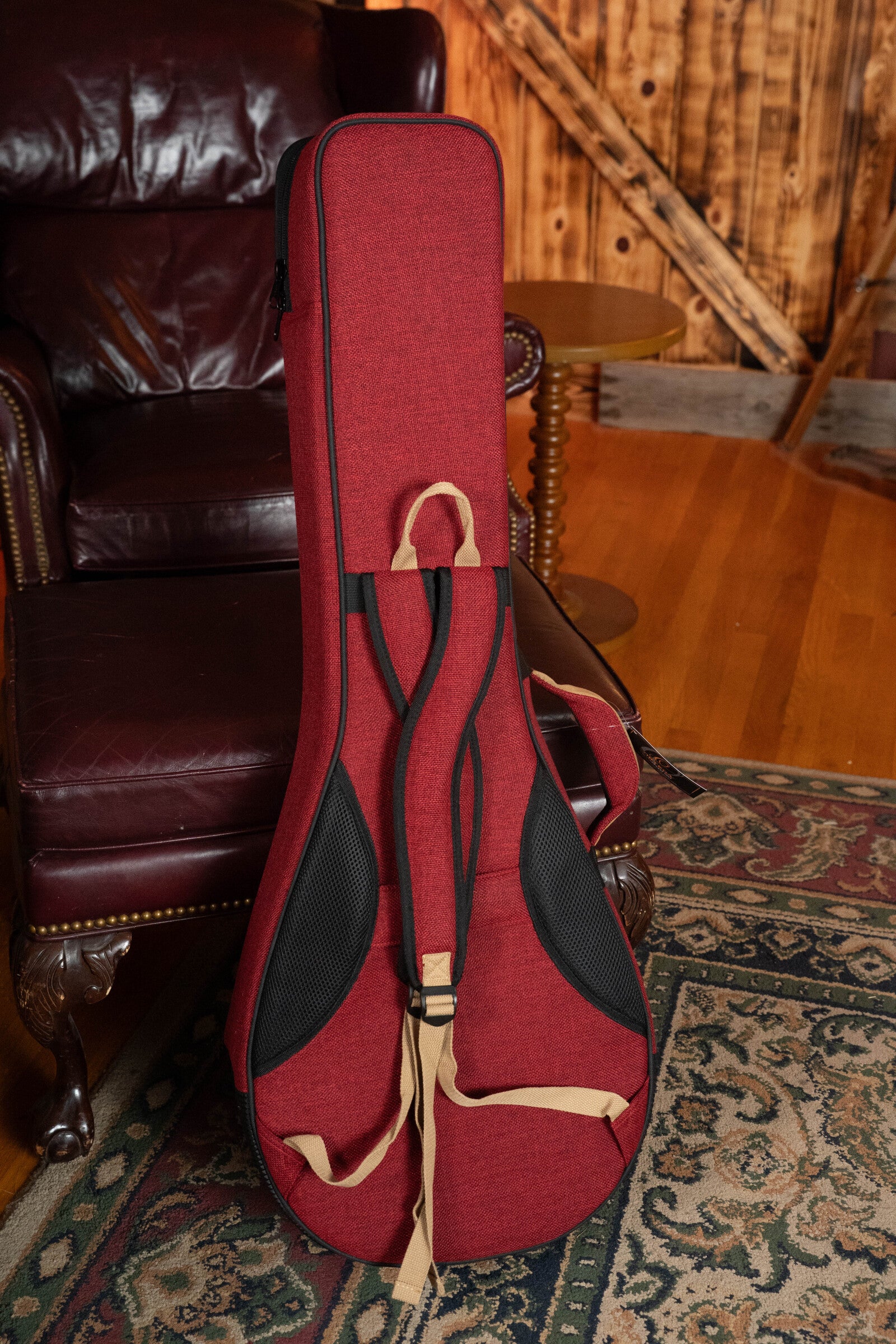 An Ortega OSOCABJ-BX Banjo Soft Case in Bordeaux Wine rests upright against a dark leather chair on a patterned rug, while another instrument case leans nearby on a wooden floor with a rustic wooden wall behind.