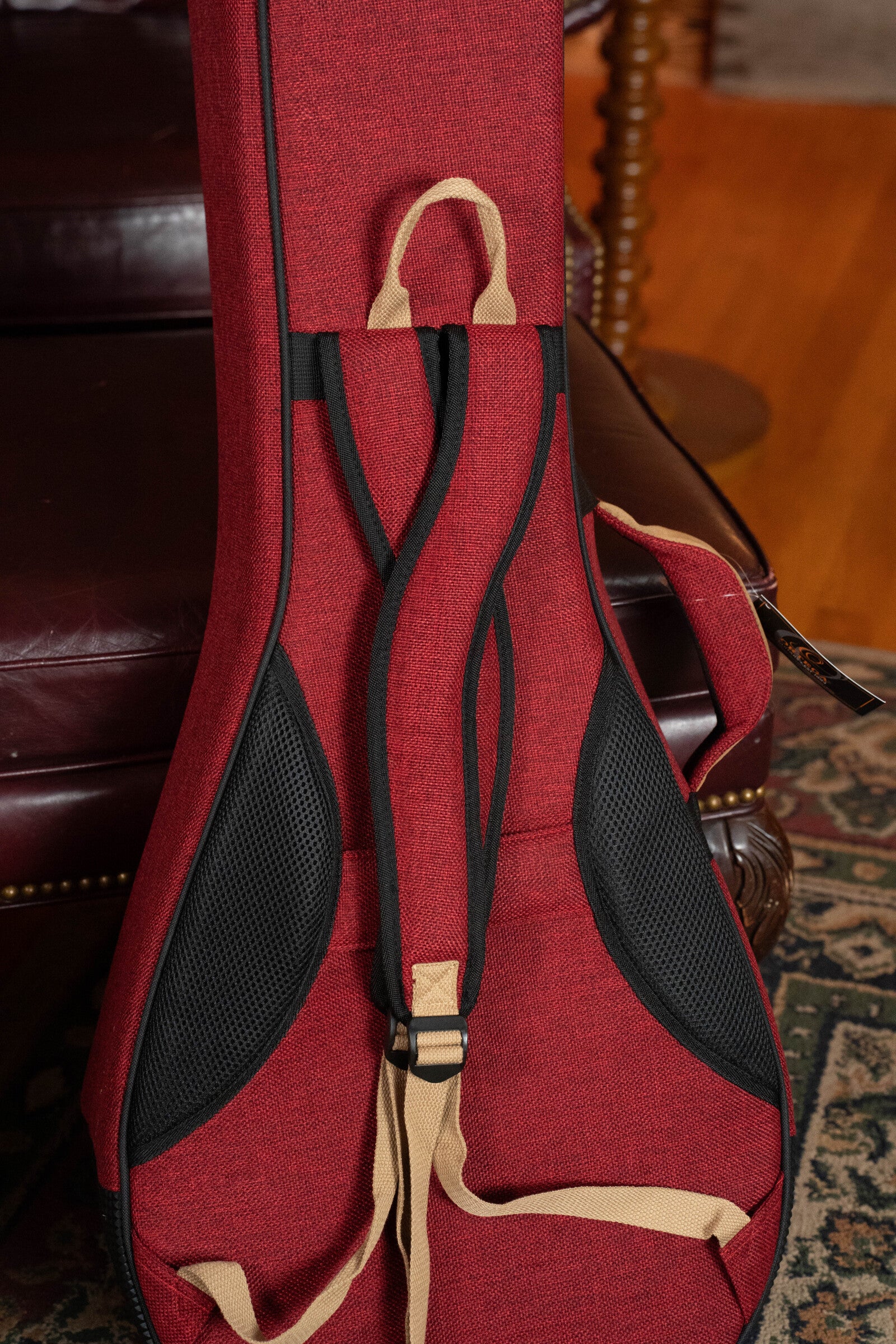 An Ortega OSOCABJ-BX Banjo Soft Case in Bordeaux Wine rests upright against a dark leather chair. The soft case features beige handles and black accents, sitting on a patterned rug with a wooden floor in the background.