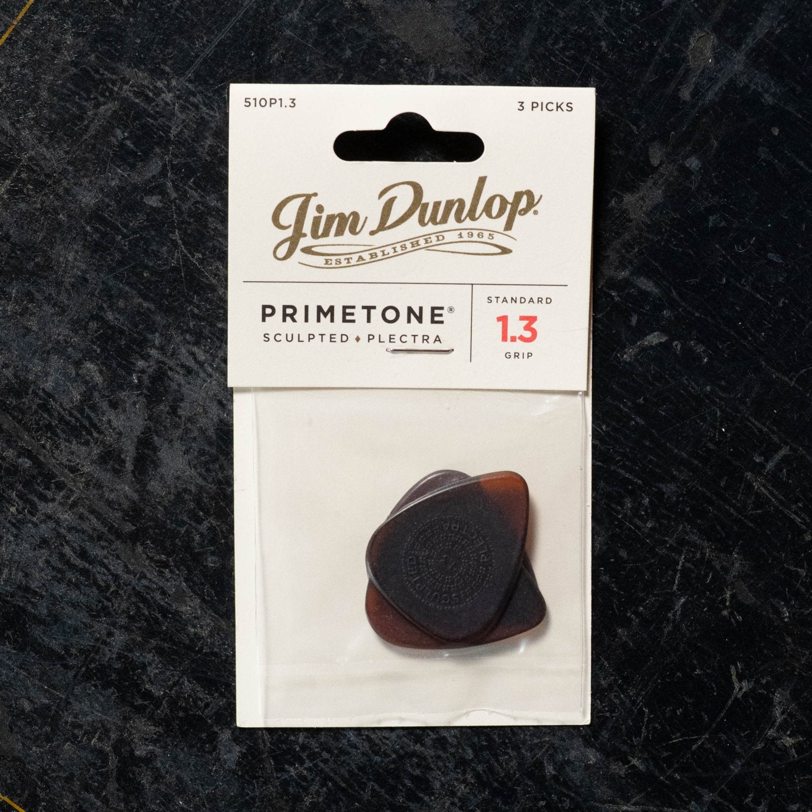 A Dunlop Primetone Standard Grip Pick 3 Pack (510P), shown in brown picks with visible grip through clear packaging, on a dark textured surface.