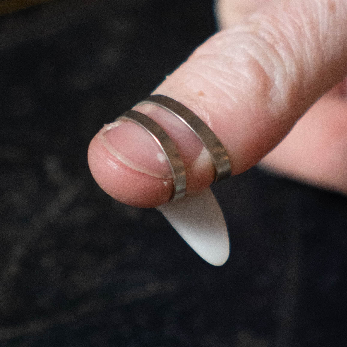 A close-up of a finger wearing two metal rings and holding the ProPik Metal/Plastic Thumb Pick Large (11-206-3002) by ProPik, featuring a white non-slip design between the rings and fingertip.