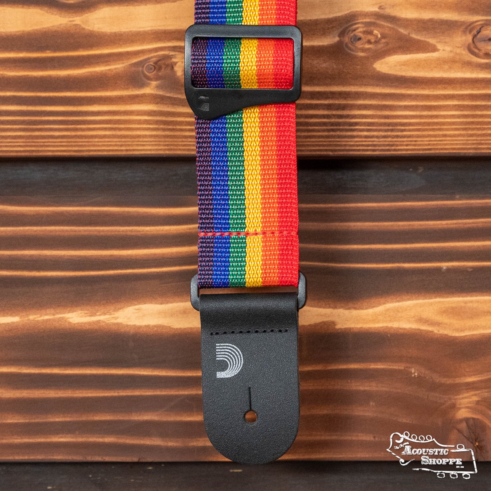 The DAddario Polypropylene Guitar Strap, Rainbow, is a heavy-duty strap with colorful stripes and a black leather end featuring the DAddario logo, displayed on a wooden background. The Acoustic Shoppe logo appears at the bottom right corner.