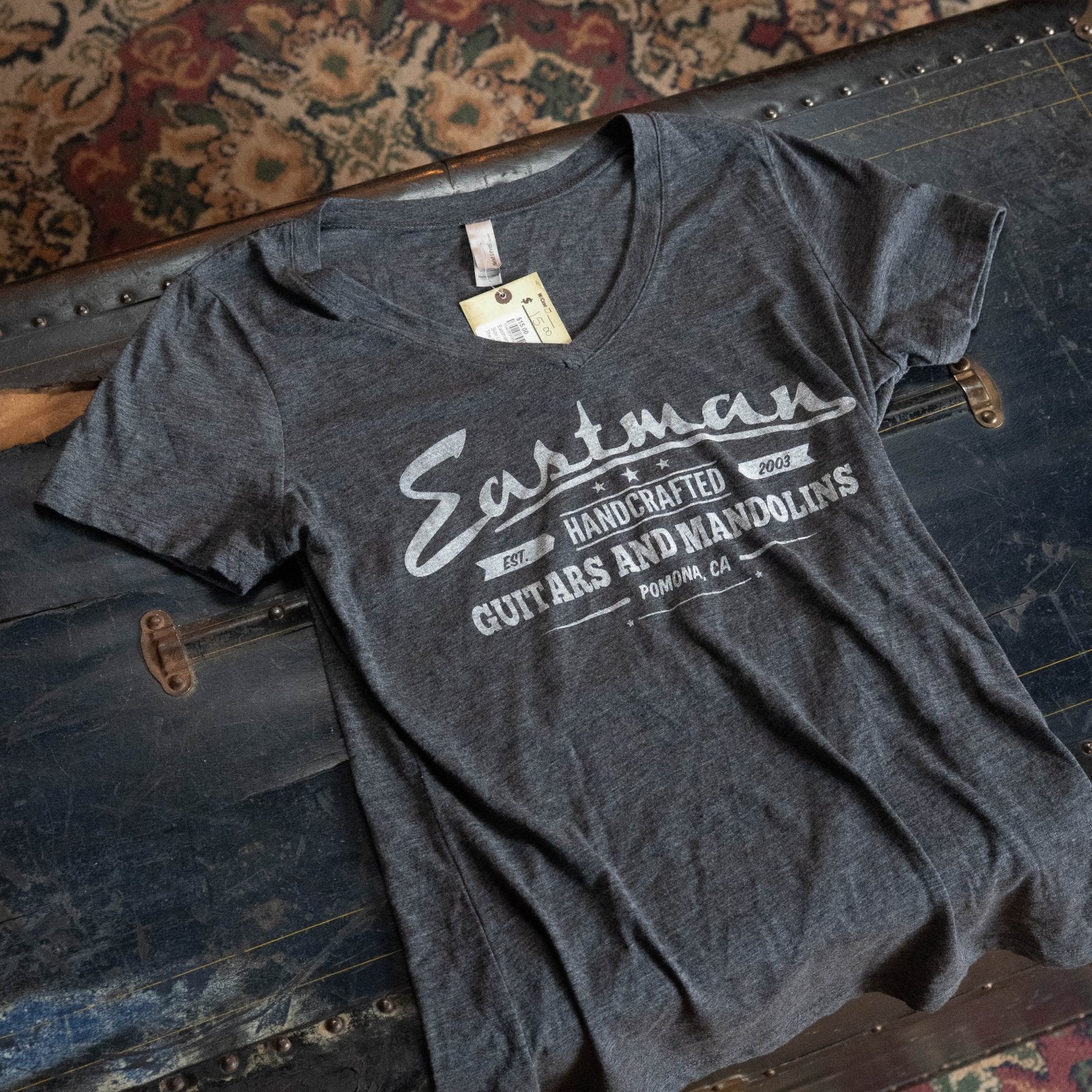 An Eastman Women's V-neck T-Shirt - Grey by The Acoustic Shoppe with white print is unfolded on a vintage trunk, while a patterned rug can be seen partially in the background.
