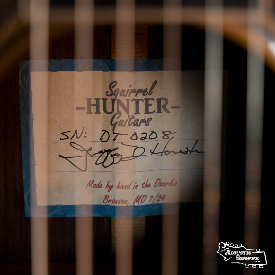 Close-up of a Squirrel Hunter Ornate Rosewood Dreadnought label inside the handmade guitar, showing serial DT-8208, a signature, and “Handcrafted in Branson, MO.” Guitar strings from the Squirrel Hunter brand are visible in front.