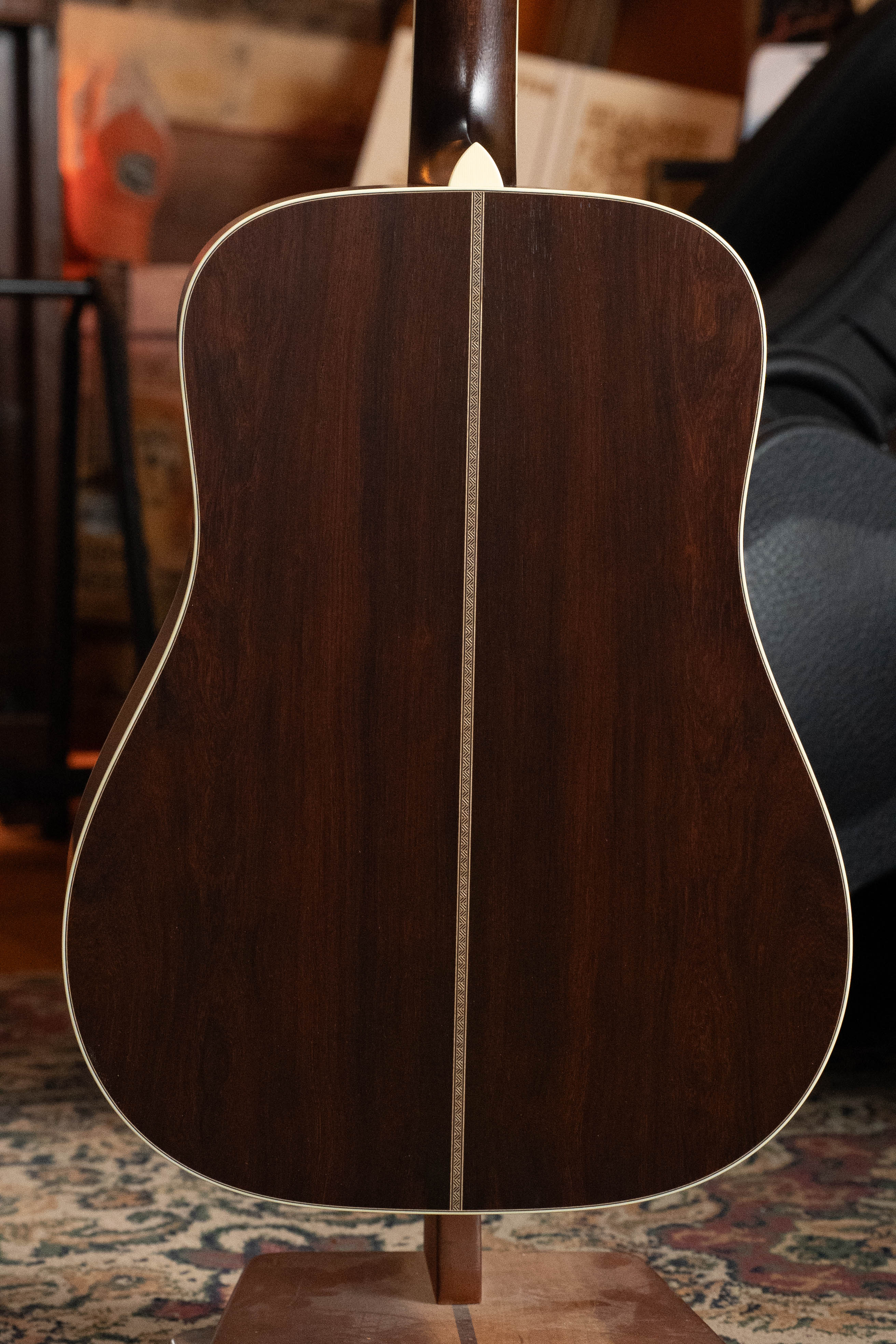 The back of a Bourgeois TAS LSH Professional Series Satin Dreadnought Guitar, featuring dark wood with a light center strip, is shown on a stand indoors. Carpet and furniture are visible in the background. *Pre-Order 25% Deposit ONLY*.