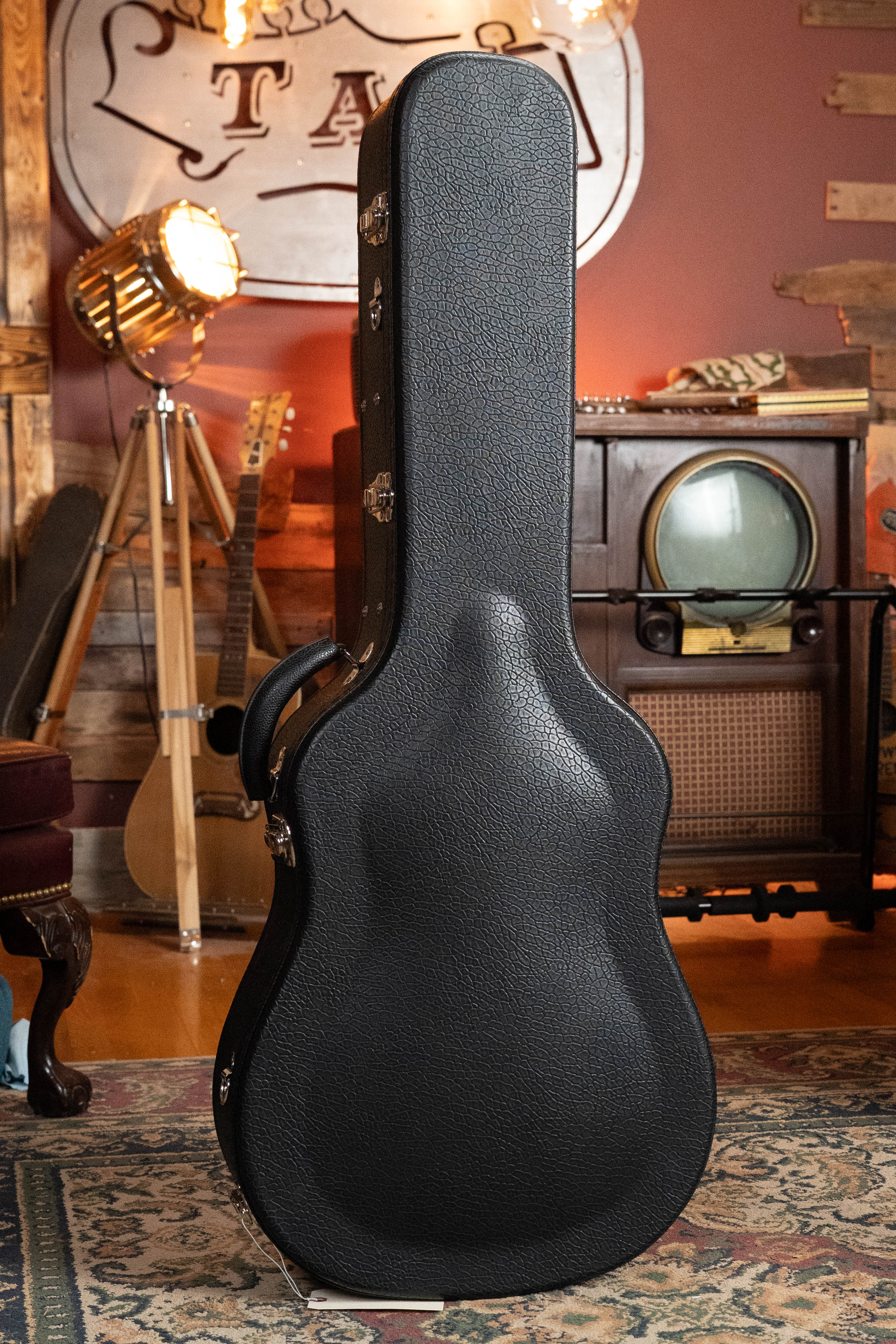 A black, hard-shell guitar case stands on a patterned rug in a cozy, vintage-inspired room—setting the scene for the *Pre-Order 25% Deposit ONLY* Bourgeois TAS LSH Professional Series Satin Dreadnought Guitar by Bourgeois.