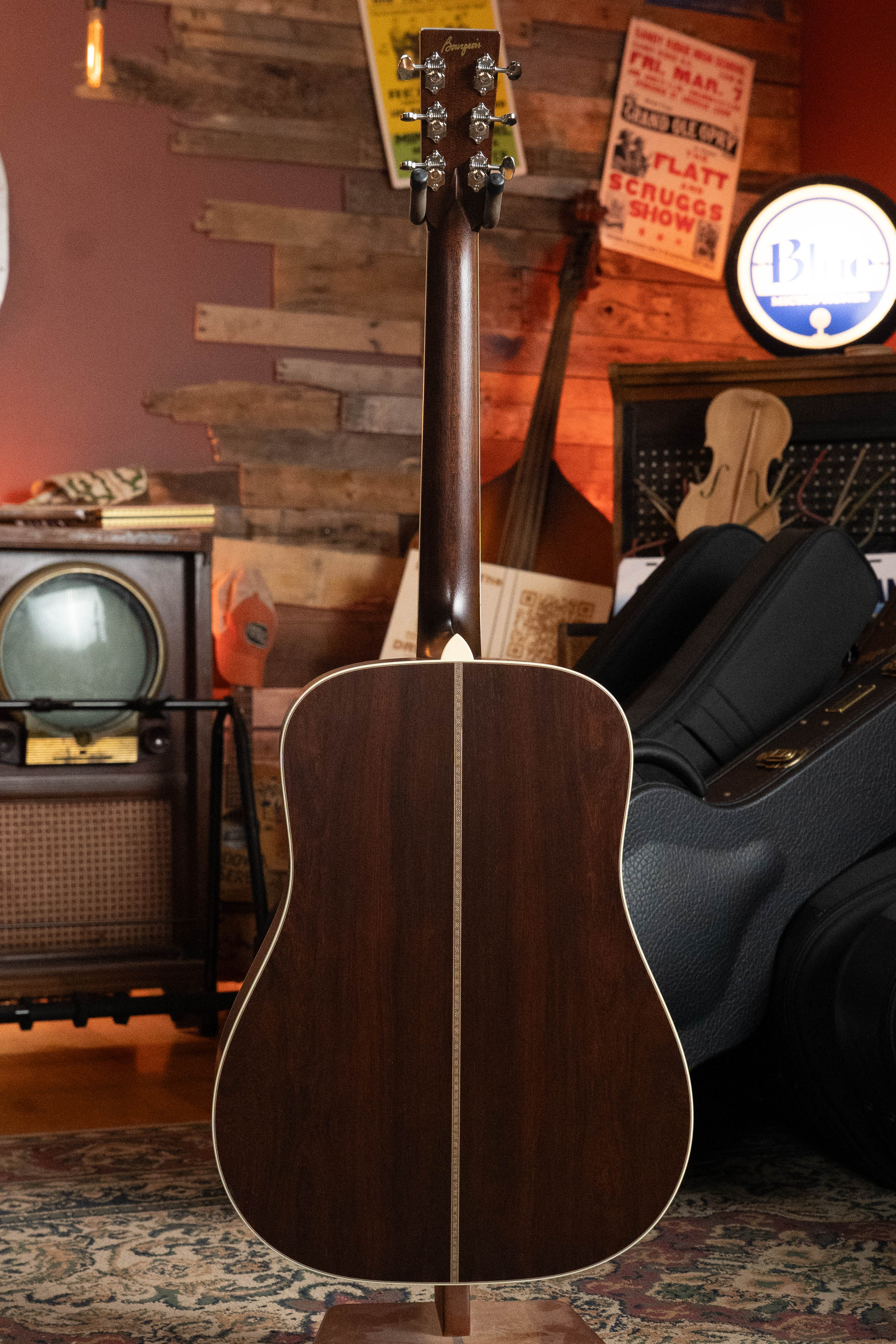A Bourgeois TAS LSH Professional Series Satin Dreadnought Guitar (*Pre-Order 25% Deposit ONLY*), with a dark wood finish, stands upright in a cozy, vintage-style room with musical instruments, a retro TV, and music posters in the background.