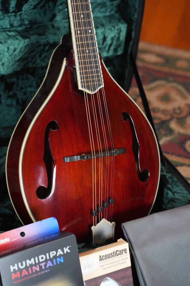 A close-up of the *TAS Exclusive* Bourgeois M5-A-CR Cordovan Red A-Style Mandolin #2121 by Bourgeois, shown in its open case with accessories like a gray cloth, Humidipak packet, and an AcoustiCare product box.