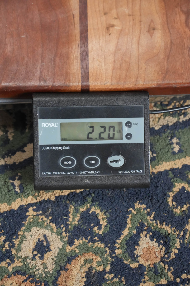 A Bourgeois *TAS Exclusive* M5-A-CR Cordovan Red A-Style Mandolin #2121 rests on a patterned carpet atop a digital shipping scale reading 2.20 pounds, with the scale partially beneath a wooden surface.