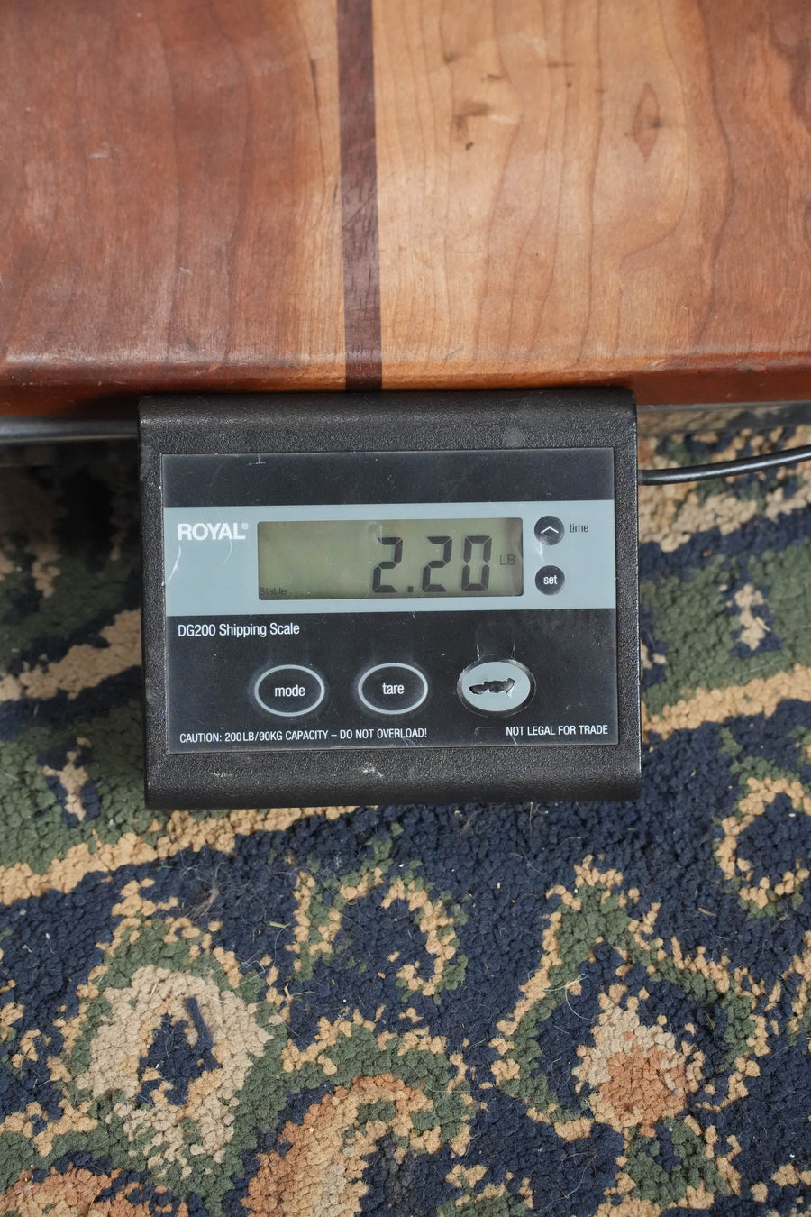 A Bourgeois *TAS Exclusive* M5-A-CR Cordovan Red A-Style Mandolin #2121 rests on a patterned carpet atop a digital shipping scale reading 2.20 pounds, with the scale partially beneath a wooden surface.
