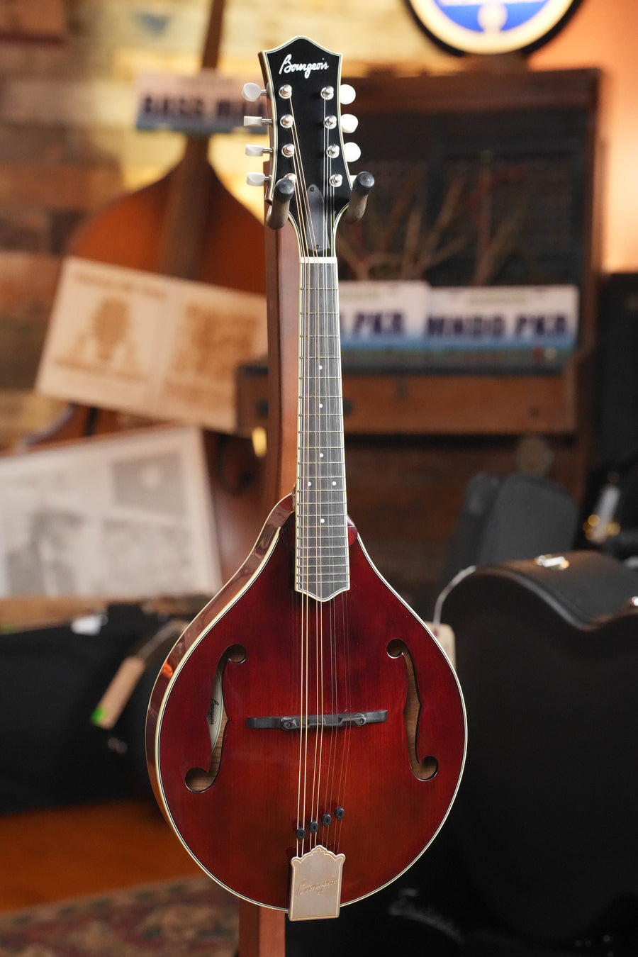 A *TAS Exclusive* Bourgeois M5-A-CR Cordovan Red A-Style Mandolin #2121 with eight tuning pegs and f-holes stands on a stand in a cozy room filled with musical equipment and instruments.