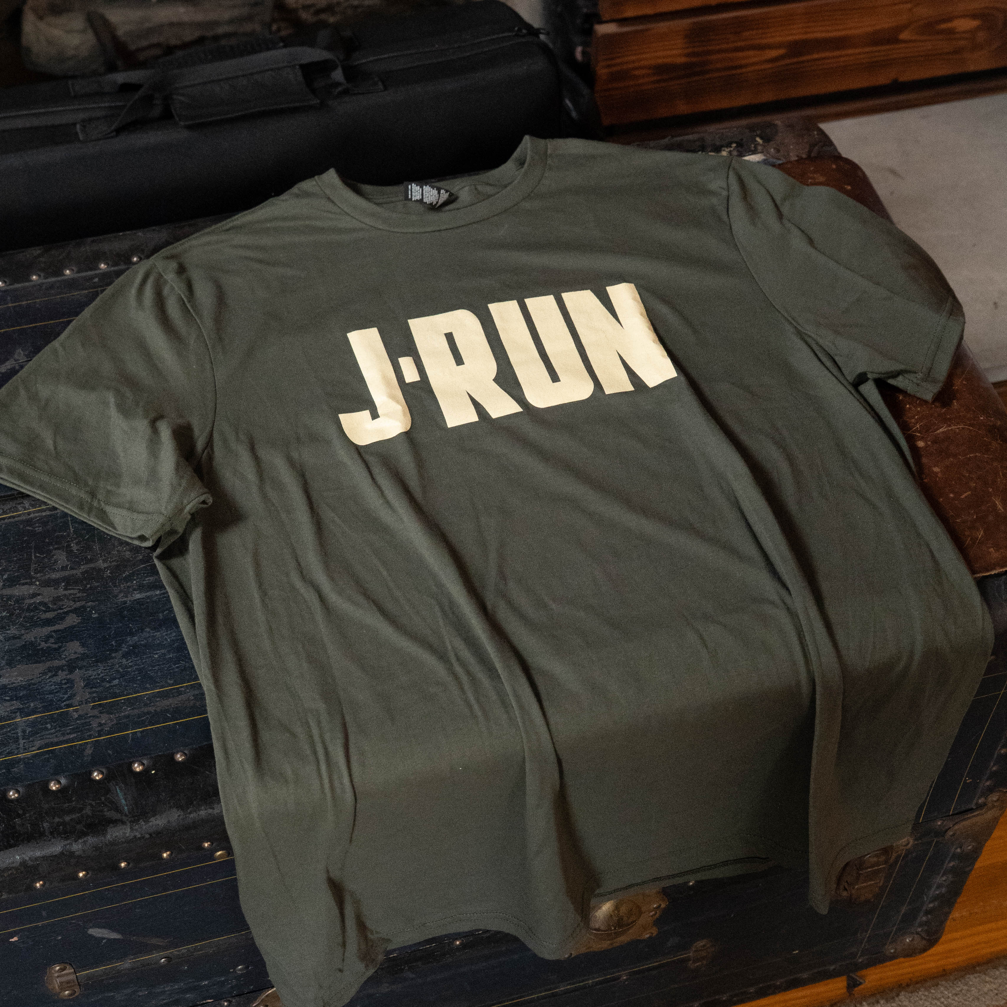 The TAS J-Run Olive Green Shirt w/Double Emblems by Amplify Screen Printing & Promotions, showcasing the bold white J-Run graphic, is displayed flat on a dark vintage trunk—a rare Acoustic Shoppe find.