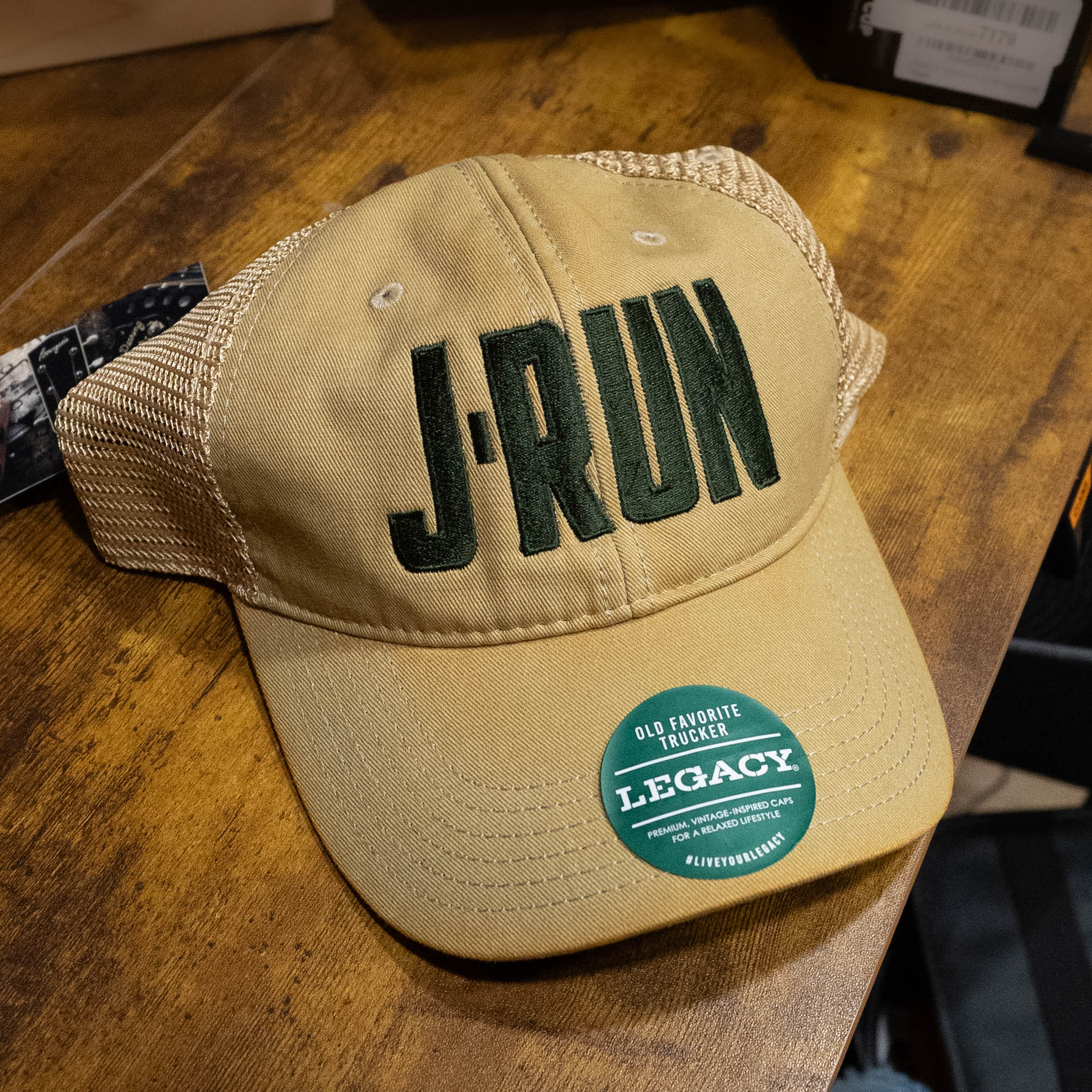 The TAS J-Run Legacy Khaki Mesh Hat w/Double Printed Emblems by Amplify Screen Printing & Promotions features bold “J-RUN” embroidery, a green “Legacy” brim sticker, and is available at The Acoustic Shoppe.