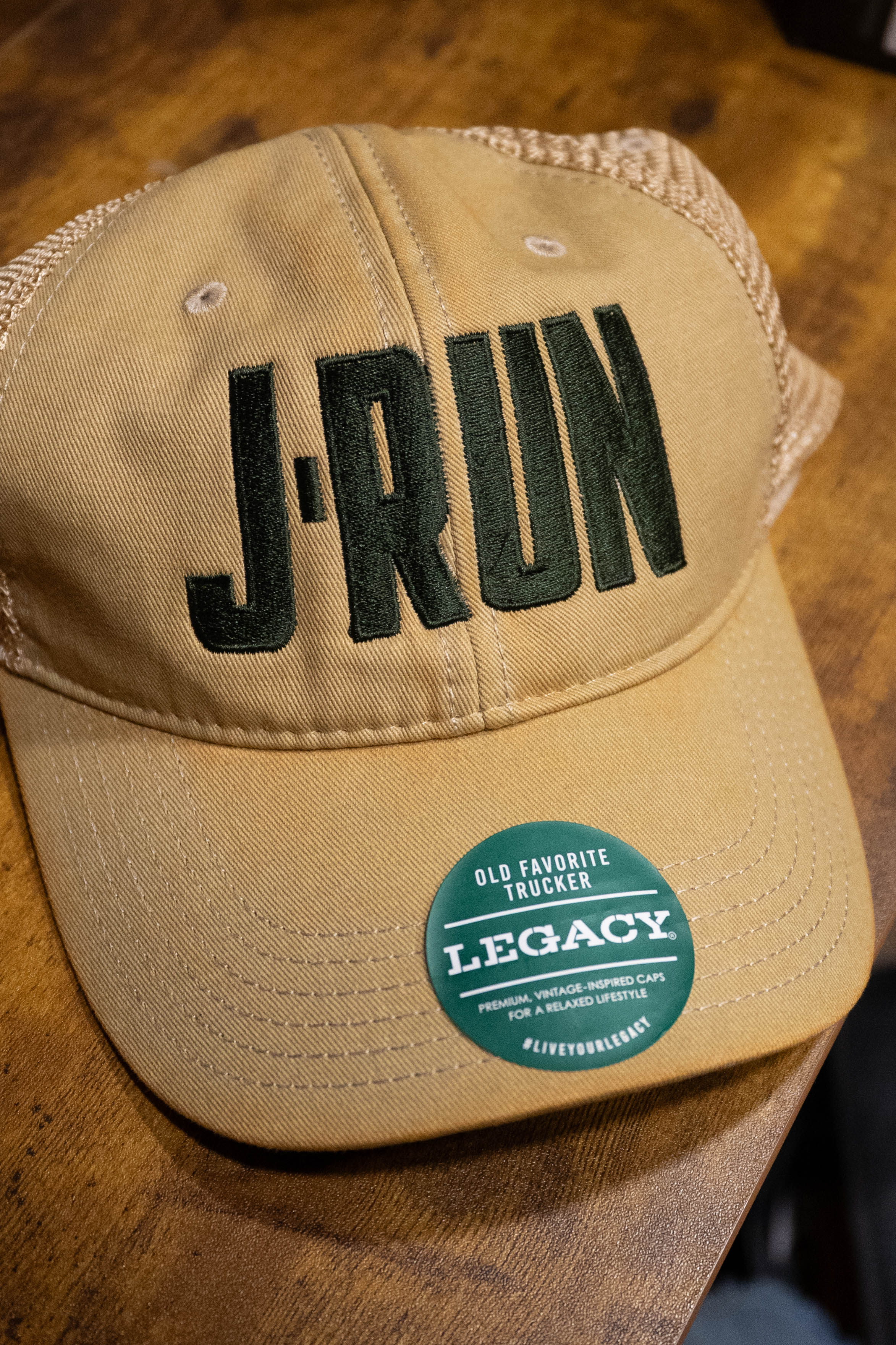 The TAS J-Run Legacy Khaki Mesh Hat w/Double Printed Emblems by Amplify Screen Printing & Promotions features bold dark green "J-RUN" text and an OLD FAVORITE TRUCKER LEGACY sticker. Available now at Acoustic Shoppe.