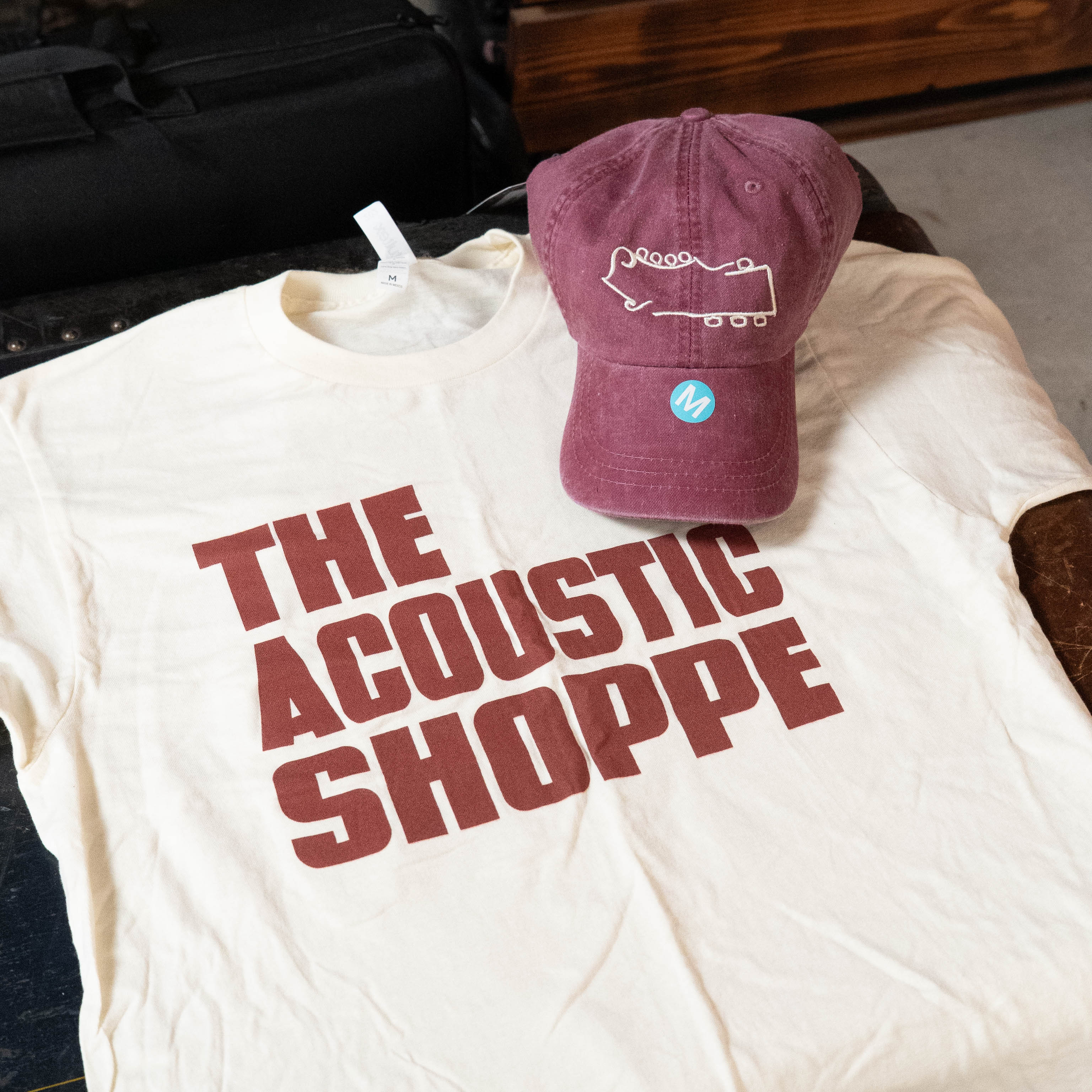 The TAS Logo Shirt and Hat Bundle by Amplify Screen Printing & Promotions includes a cream shirt with bold red "THE ACOUSTIC SHOPPE" text and a maroon hat featuring a white guitar headstock outline in a blue circle logo.