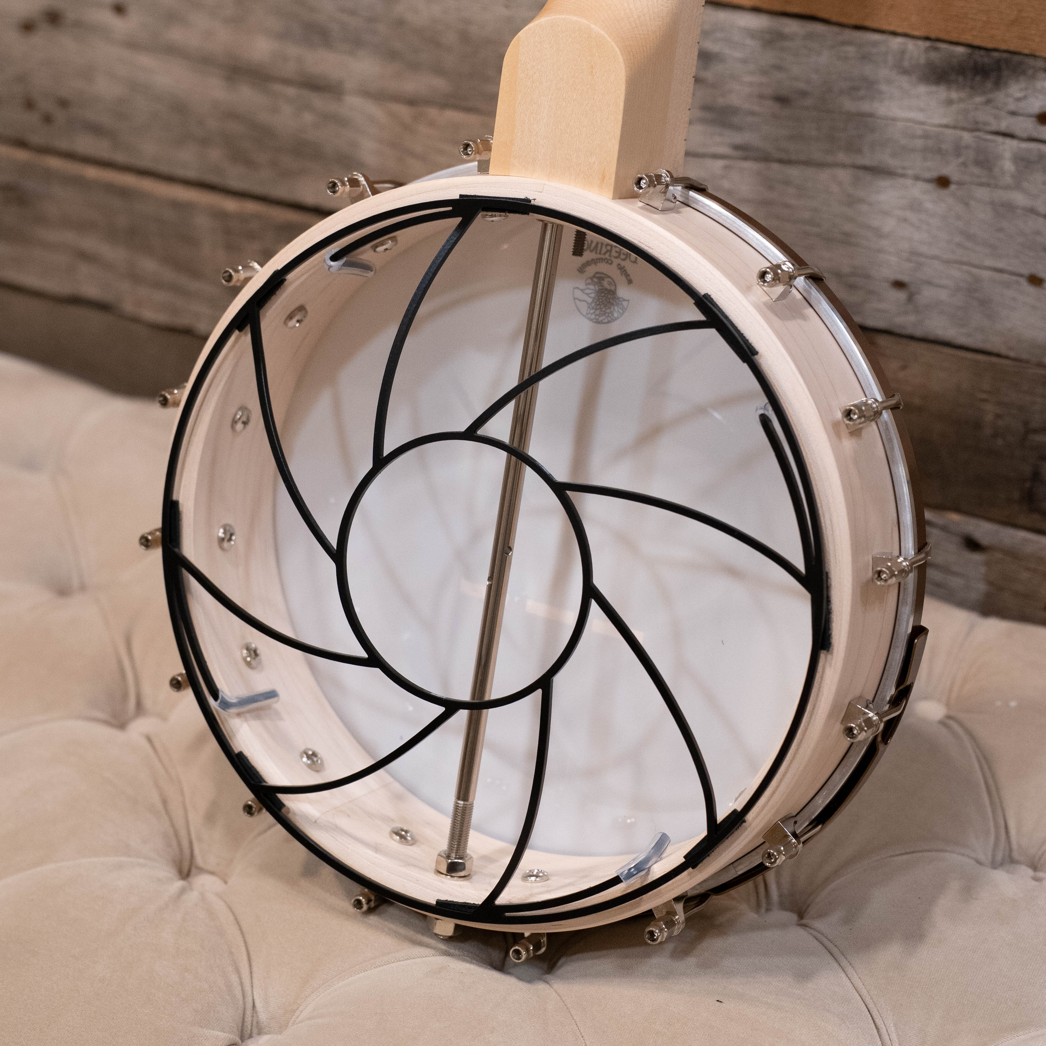 A Tone-Gard Open Back Banjo (11") by Tone-Gard, featuring a decorative black metal swirl design, rests on a beige tufted surface with its wooden frame and metal hardware clearly visible.