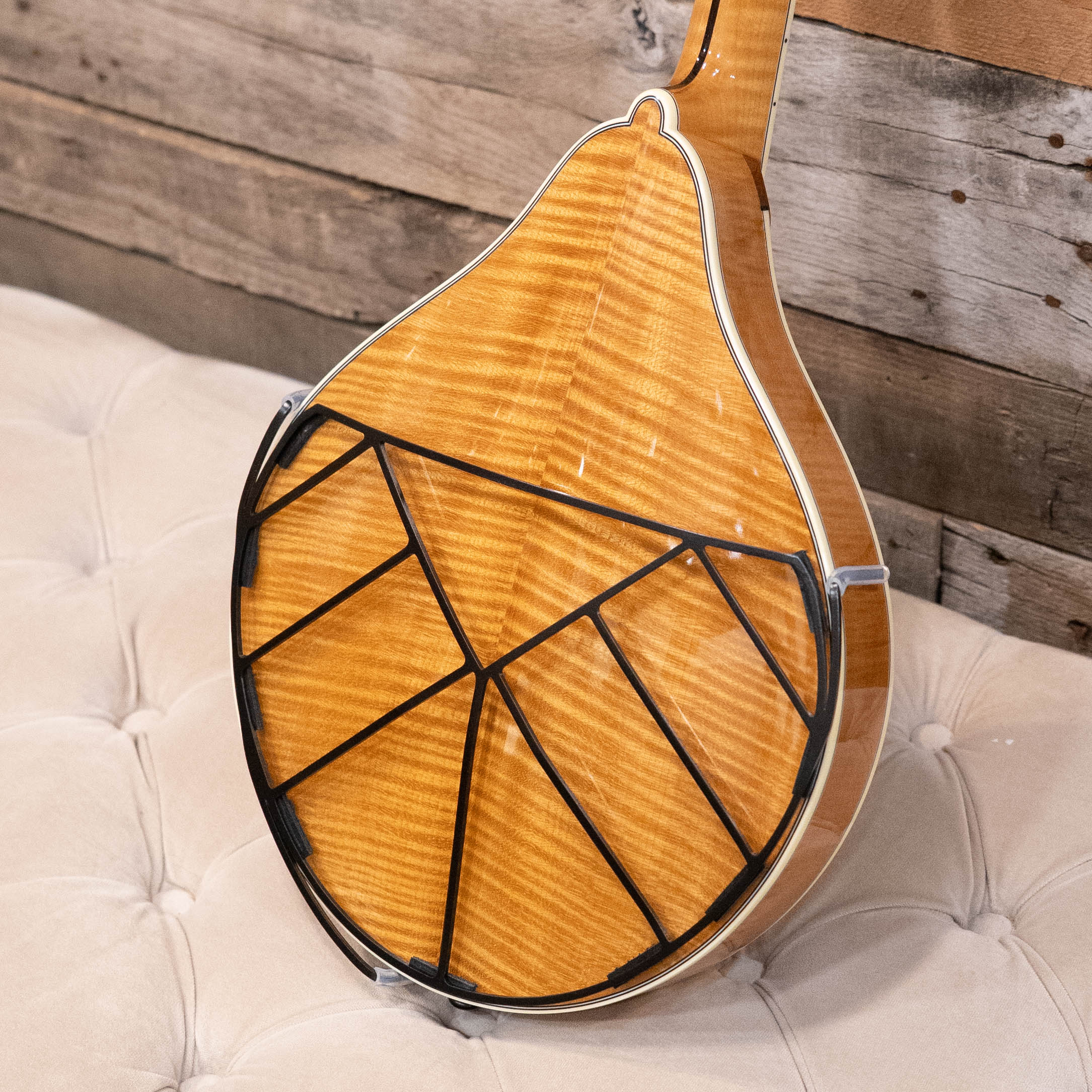 A close-up of the Tone-Gard Mandolin - Deco by Tone-Gard shows its glossy, flamed wood back and unique geometric black Tone-Gard pattern that protects the instrument, resting on a beige tufted surface with a wooden wall behind it.