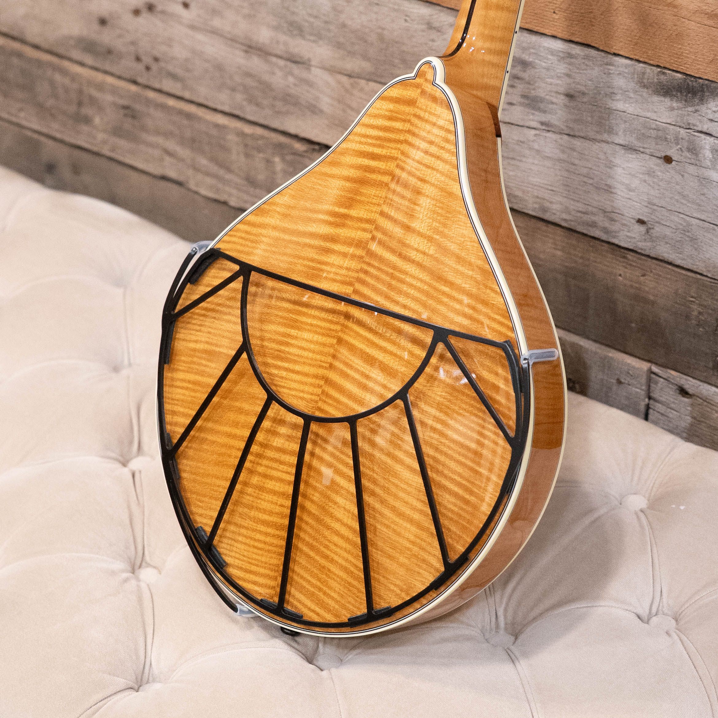 A close-up of a mandolin with a sunburst finish shows the Tone-Gard Mandolin - Sunrise back protector by Tone-Gard attached. The instrument rests on a tufted cream cushion, with a wooden wall in the background.