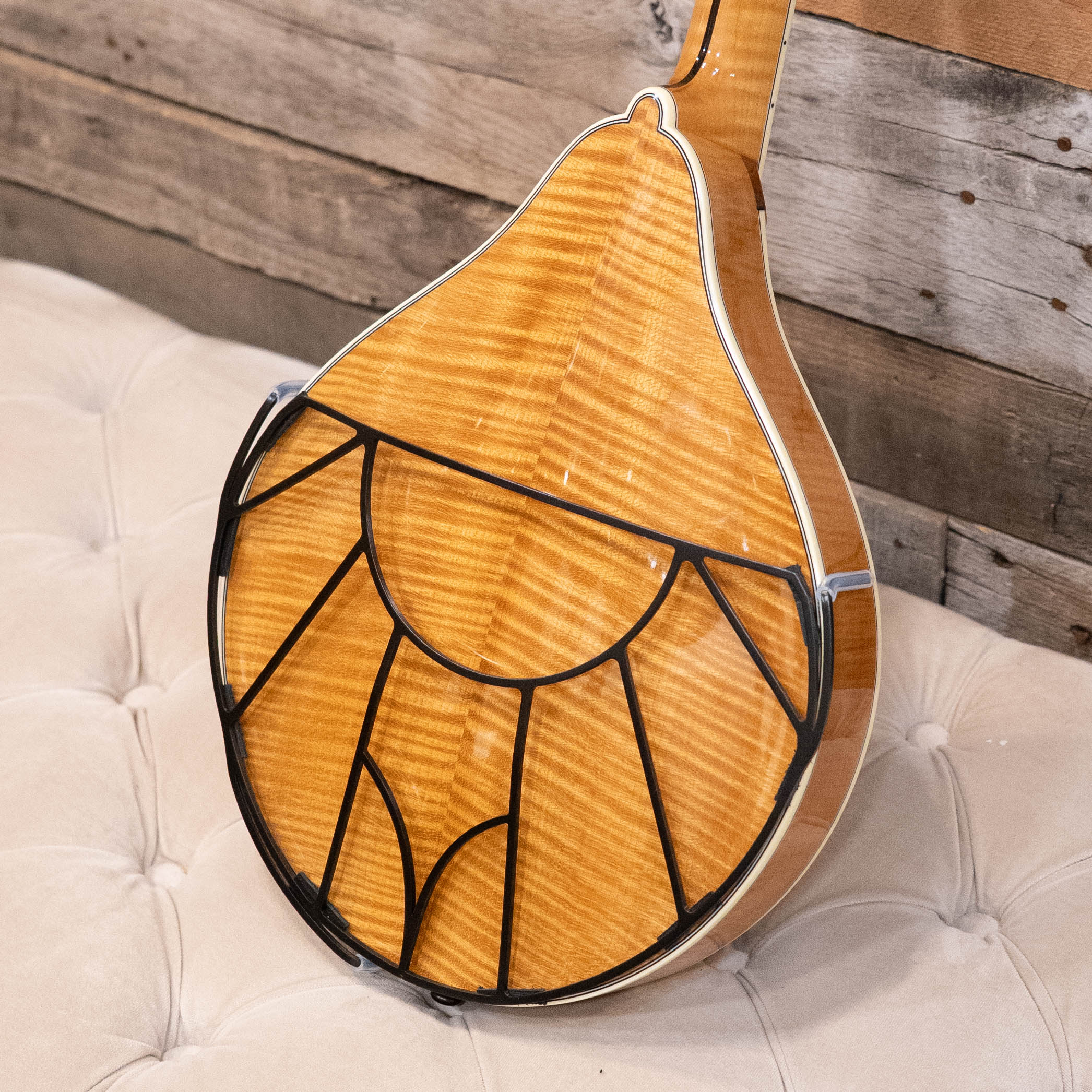 A close-up of a mandolin’s glossy, striped back features the Tone-Gard Mandolin - Vintage back protector by Tone-Gard, resting on a tufted cream cushion against a wooden wall.