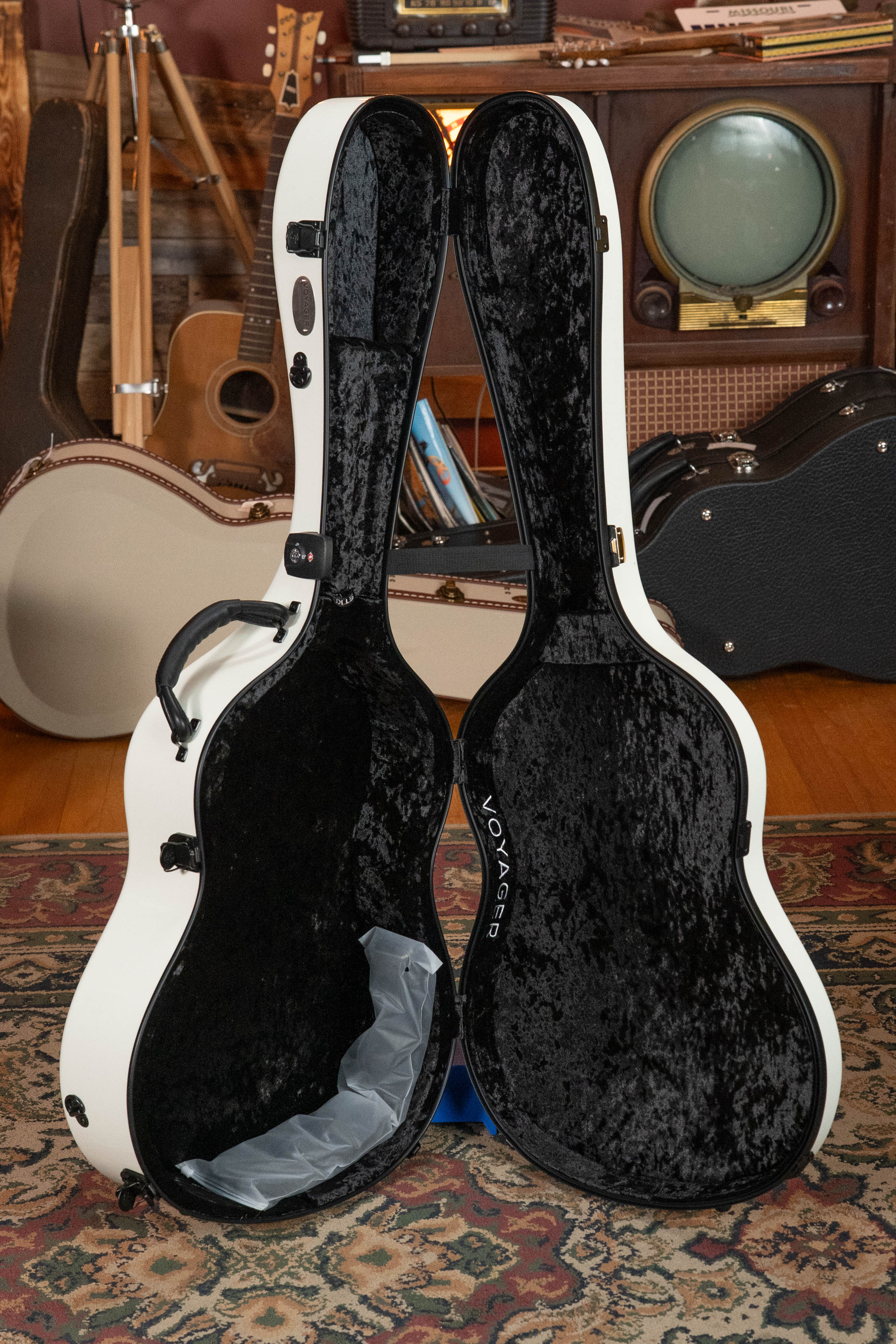 A TKL Voyager Universal Pearl Glacier/Blue Carbon Fiber Dreadnought Case with a black plush interior stands open on a rug, surrounded by guitars, cases, and vintage audio gear in a cozy room.