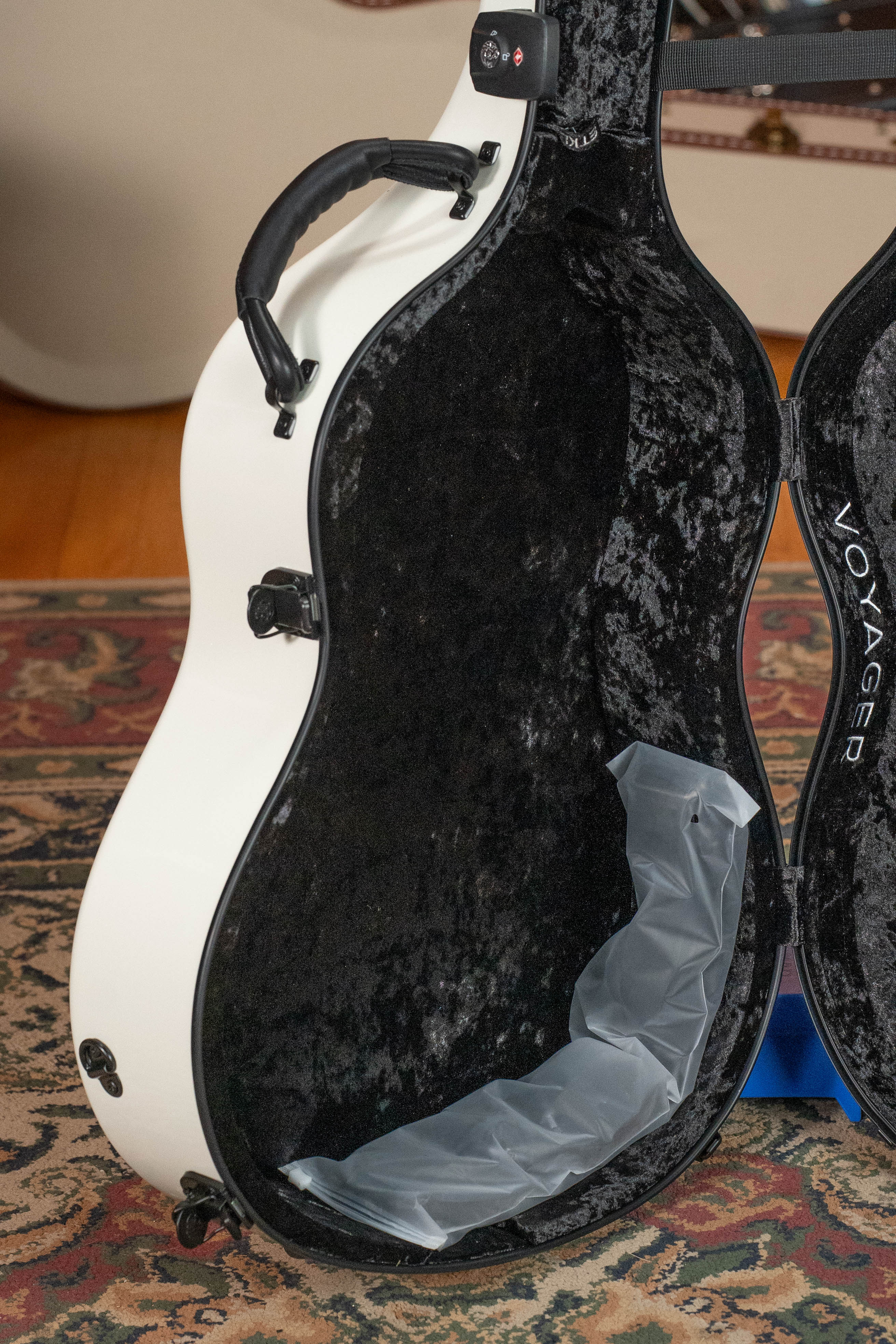 The TKL Voyager Universal Pearl Glacier/Blue Carbon Fiber Dreadnought Case, lined with black velvet and cushioning, is open to show its protective handle and packing material. This white TKL case rests on a patterned rug—perfect for touring musicians.