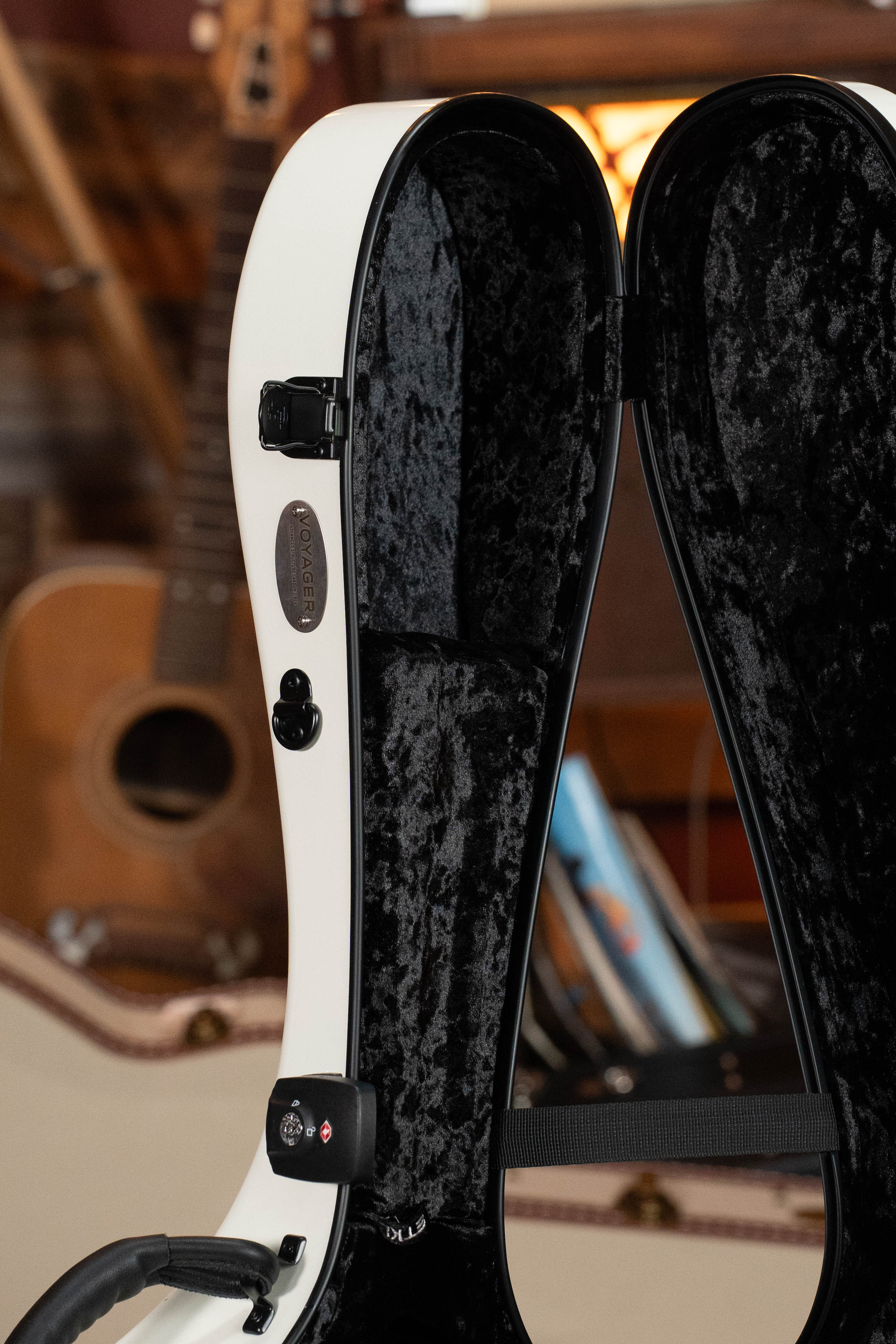 The TKL Voyager Universal Pearl Glacier/Blue Carbon Fiber Dreadnought Case stands upright, its plush black interior visible. An acoustic guitar and books in the background add a warm, cozy atmosphere.