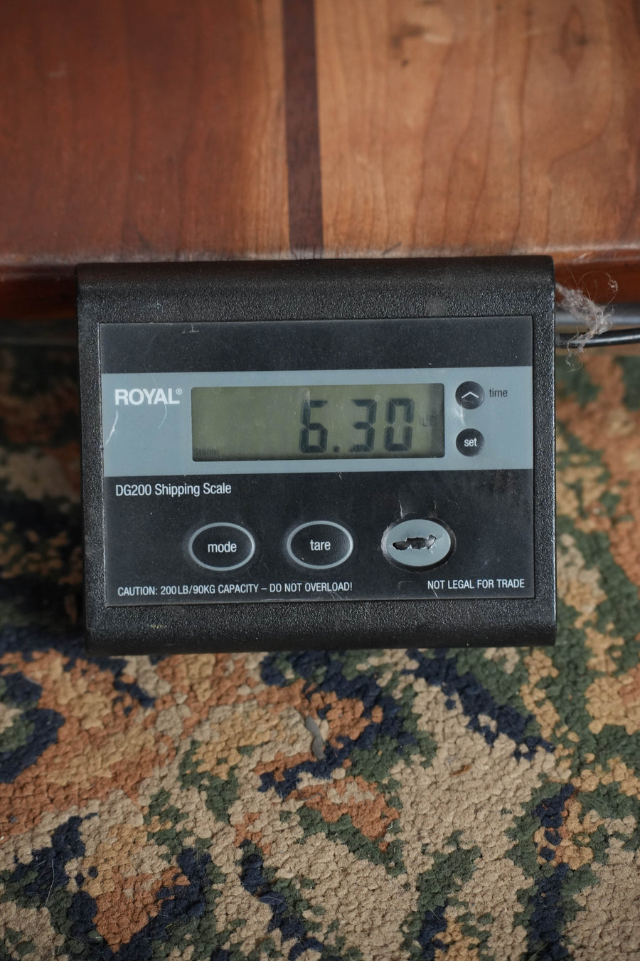 A Bacon digital shipping scale on a patterned carpet displays a (Used) 1928 Bacon Style C Maple/Mahogany Resonator Banjo #2530 weighing 6.30 pounds. The scale's mode, tare, and set buttons are visible along with brand and capacity details.
