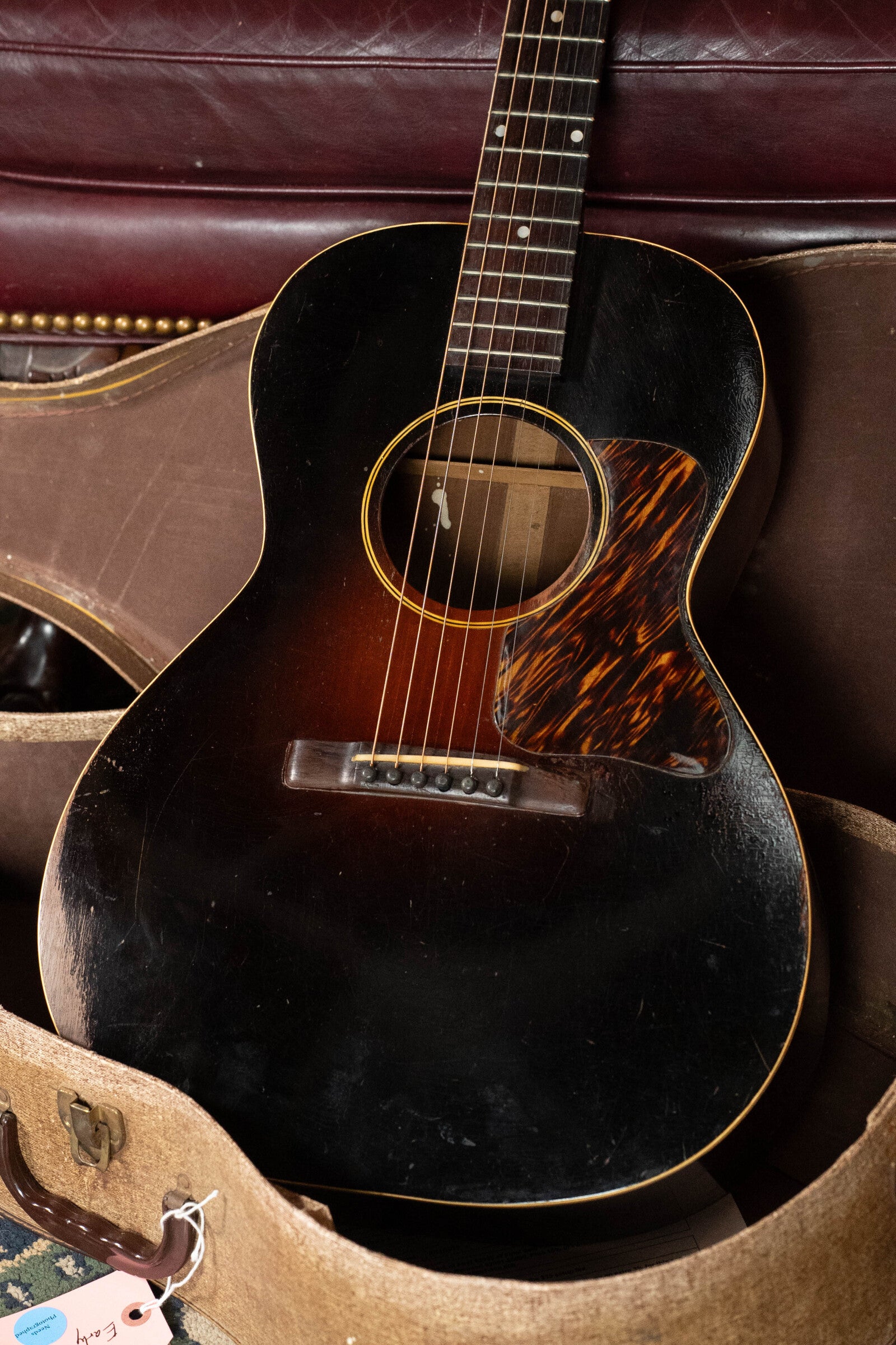 A (Used) 1933 Gibson LOO Adirondack/Mahogany OO Tobacco Sunburst Acoustic Guitar #550 by Gibson rests in an open case with a maroon leather sofa behind it, showing its tortoiseshell pickguard and vintage wear.