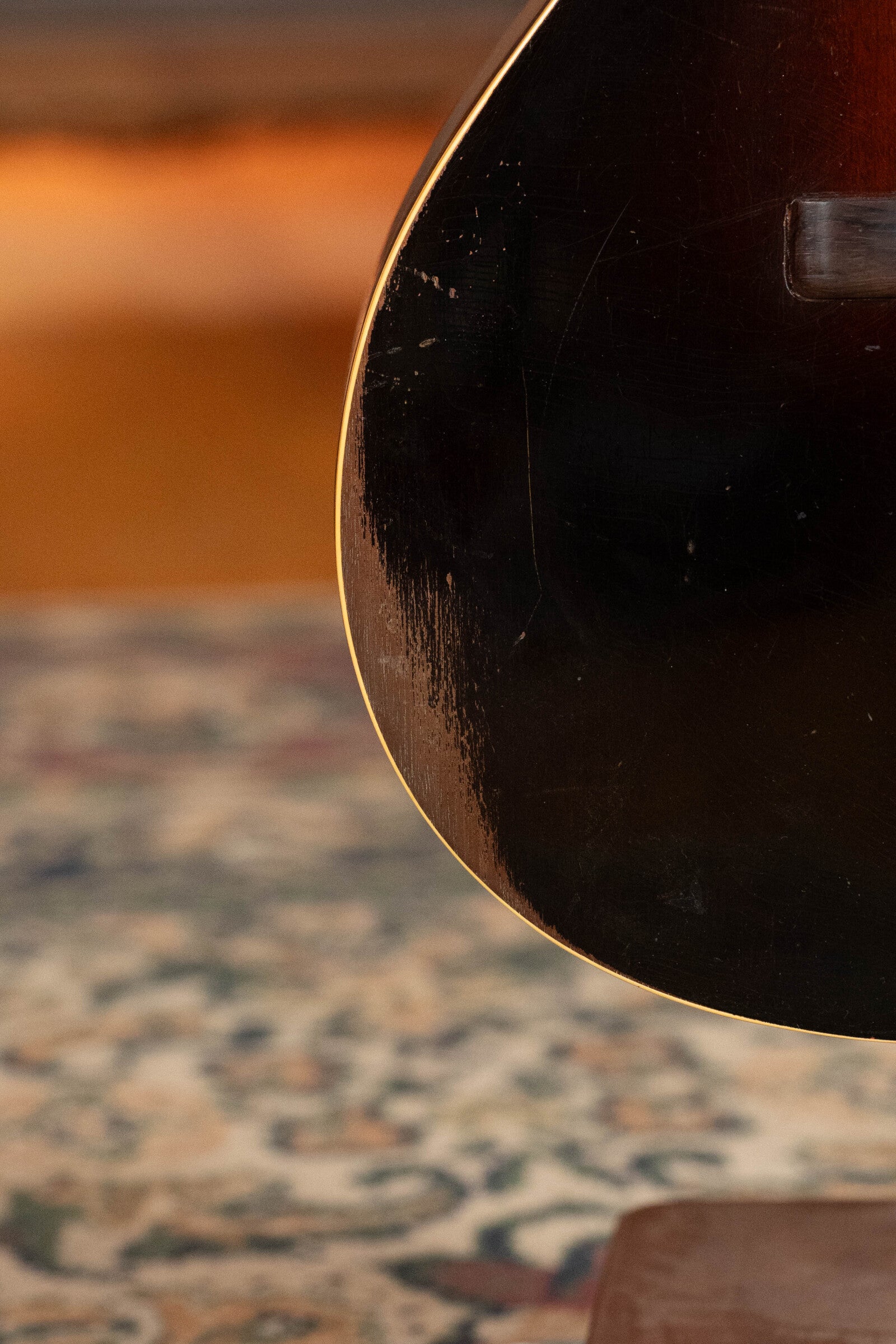 Close-up of a 1933 Gibson LOO Adirondack/Mahogany OO Tobacco Sunburst Acoustic Guitar (#550) resting on patterned carpet, showcasing its dark, glossy body under warm, blurred lighting.