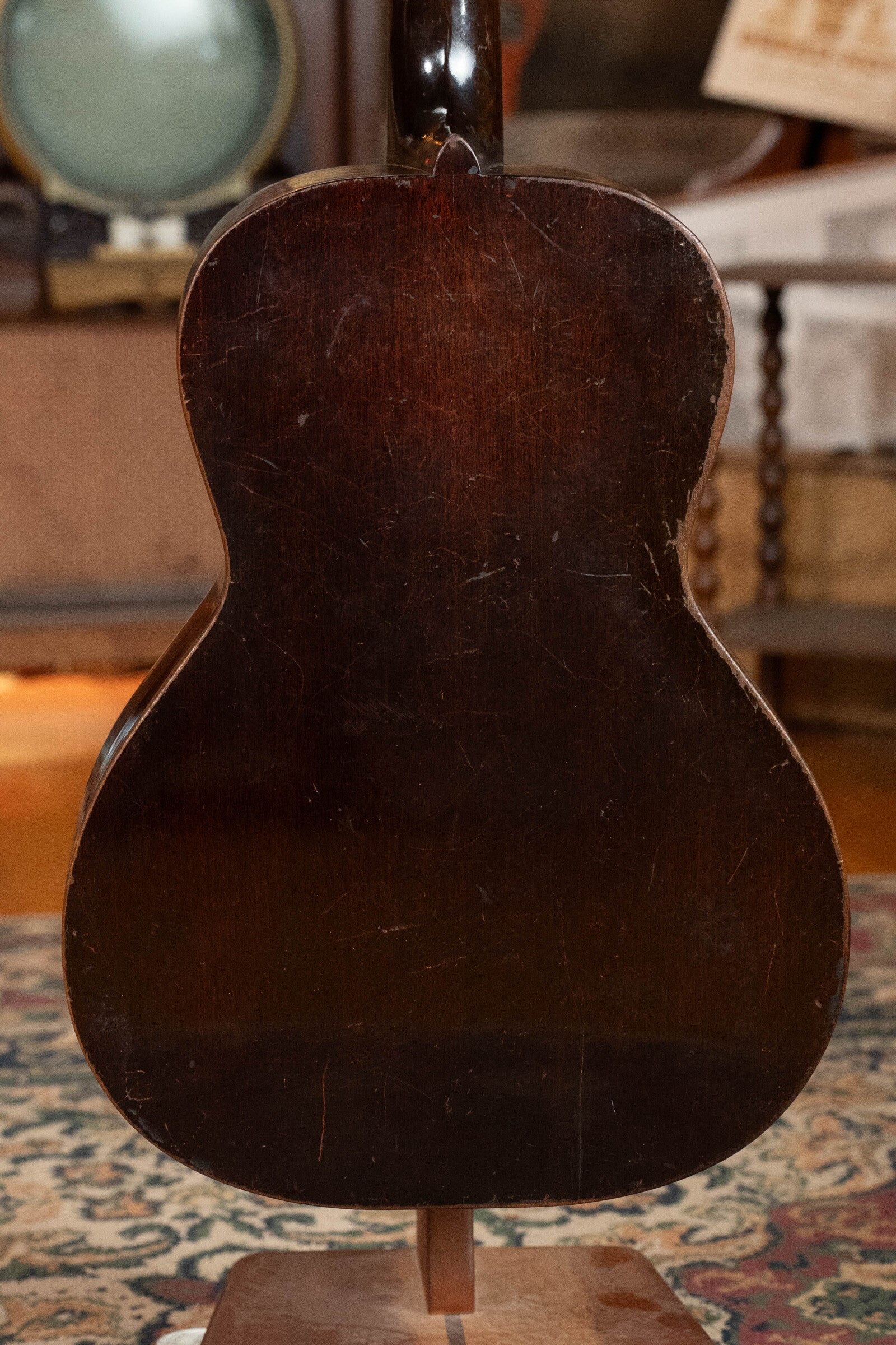The image features the back of a used 1933 Gibson LOO Adirondack/Mahogany OO Tobacco Sunburst Acoustic Guitar (#550) with visible wear, displayed on a stand indoors among vintage furniture and a patterned rug.