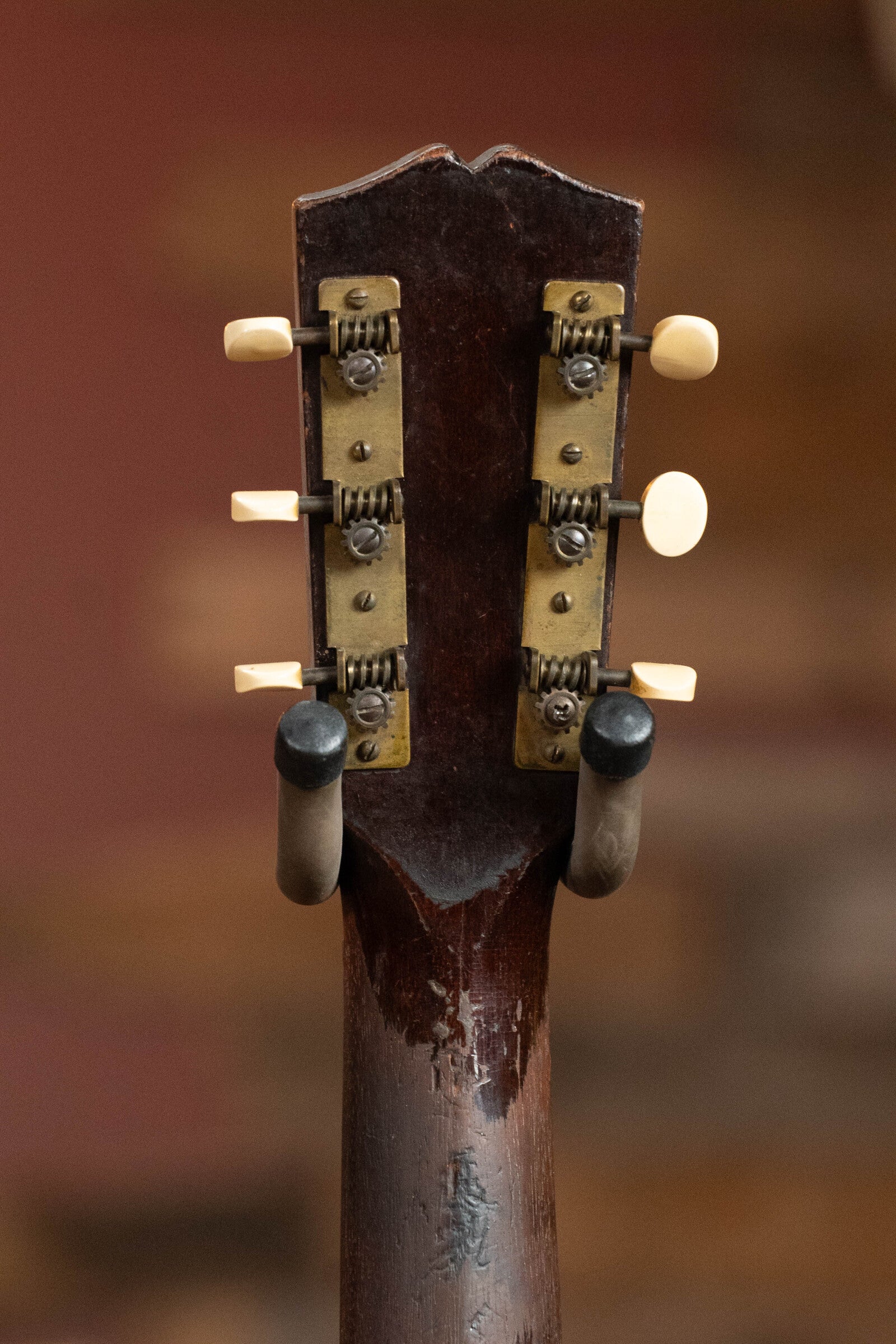 The image shows the back of a worn Gibson 1933 L-00 acoustic guitar headstock with vintage tuners and cream pegs, model #550, set against a blurred brown background.