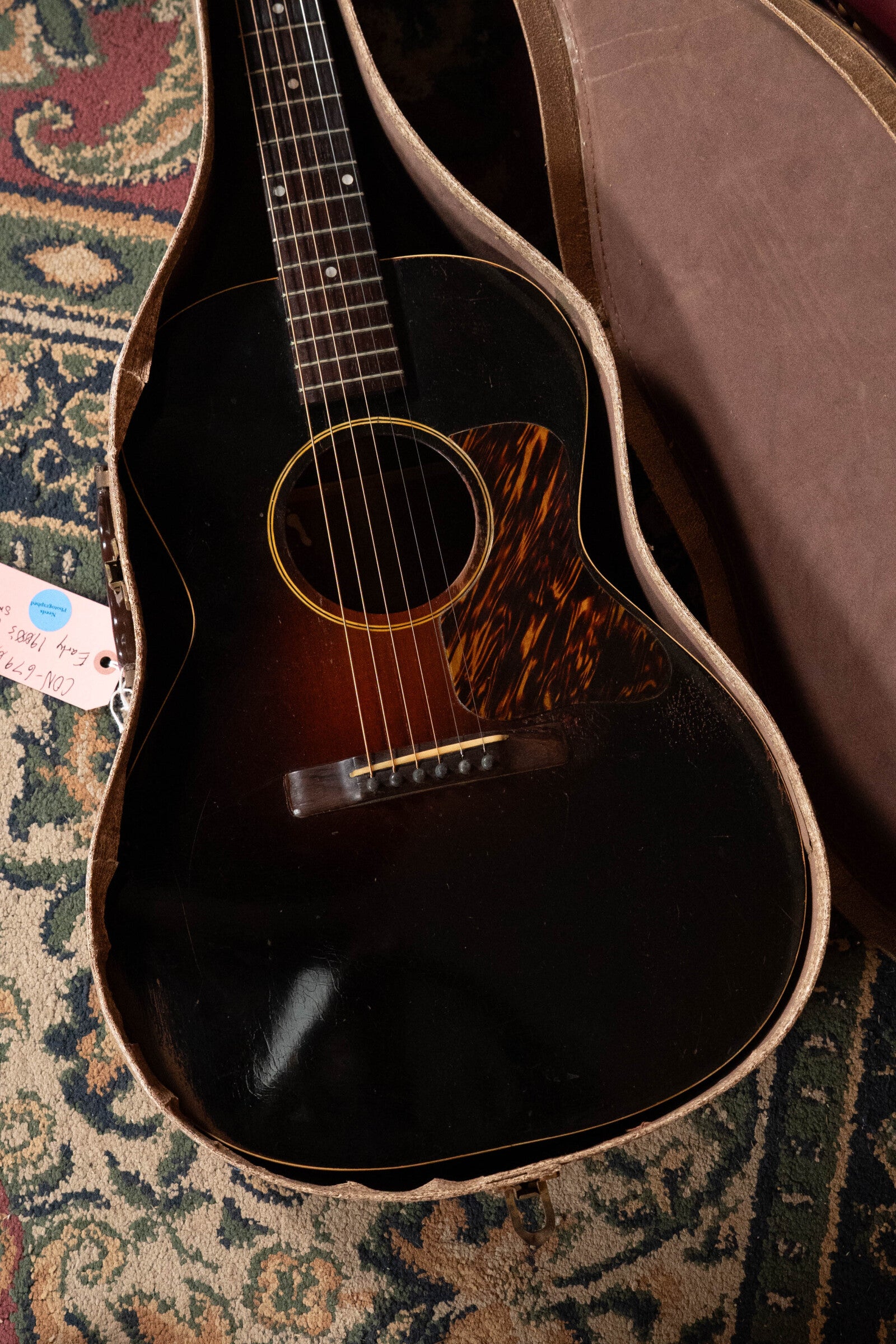 A (Used) 1933 Gibson LOO Adirondack/Mahogany OO Tobacco Sunburst Acoustic Guitar #550 by Gibson rests in an open hard case on a patterned rug, with a handwritten tag attached to the prewar guitar’s case.