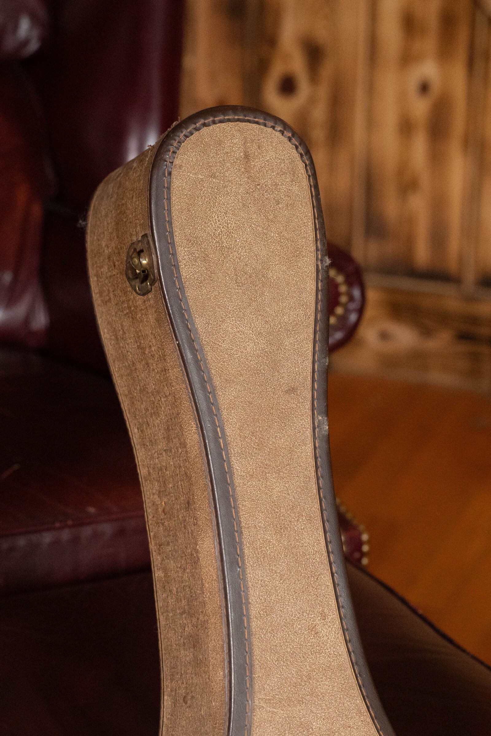 A close-up of a brown suede guitar case, possibly containing a (Used) 1933 Gibson LOO Adirondack/Mahogany OO Tobacco Sunburst Acoustic Guitar #550 by Gibson, rests on a dark red leather chair with a wooden wall and floor in the background.