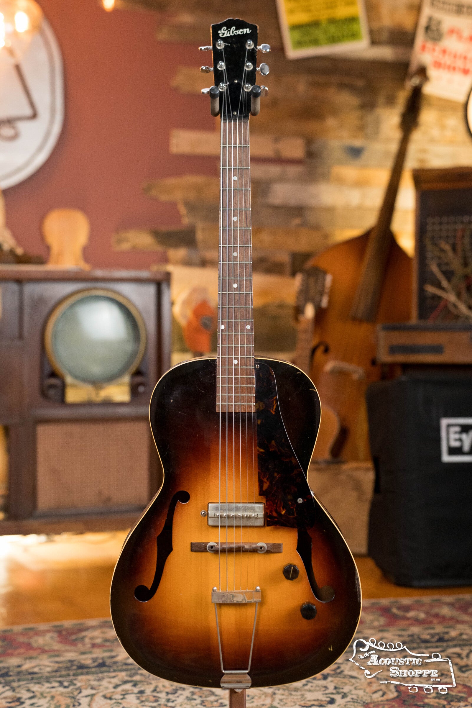 A (Used) 1942 Gibson ES-125 by Gibson, a vintage hollow-body guitar with sunburst finish and classic P-90 pickup, stands upright in a cozy, retro-themed room with warm wood accents and musical gear.