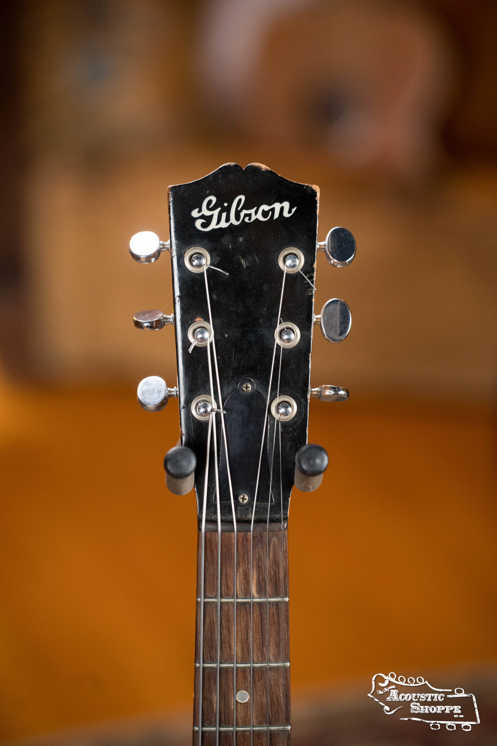 Close-up of the headstock of a (Used) 1942 Gibson ES-125 by Gibson, featuring tuning pegs, logo, and fretboard. Warm blurred background with Acoustic Shoppe logo in the bottom right corner.