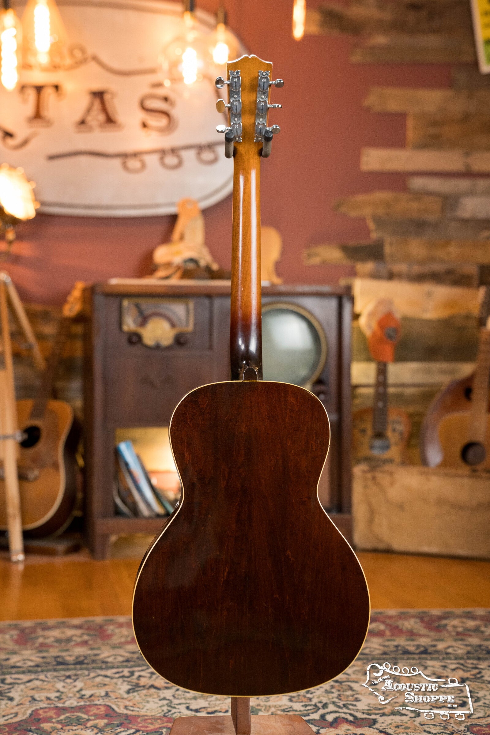 A (Used) 1942 Gibson ES-125 by Gibson is displayed from the back, standing in a cozy room with wooden accents, vintage décor, other guitars, and warm lighting in the background.