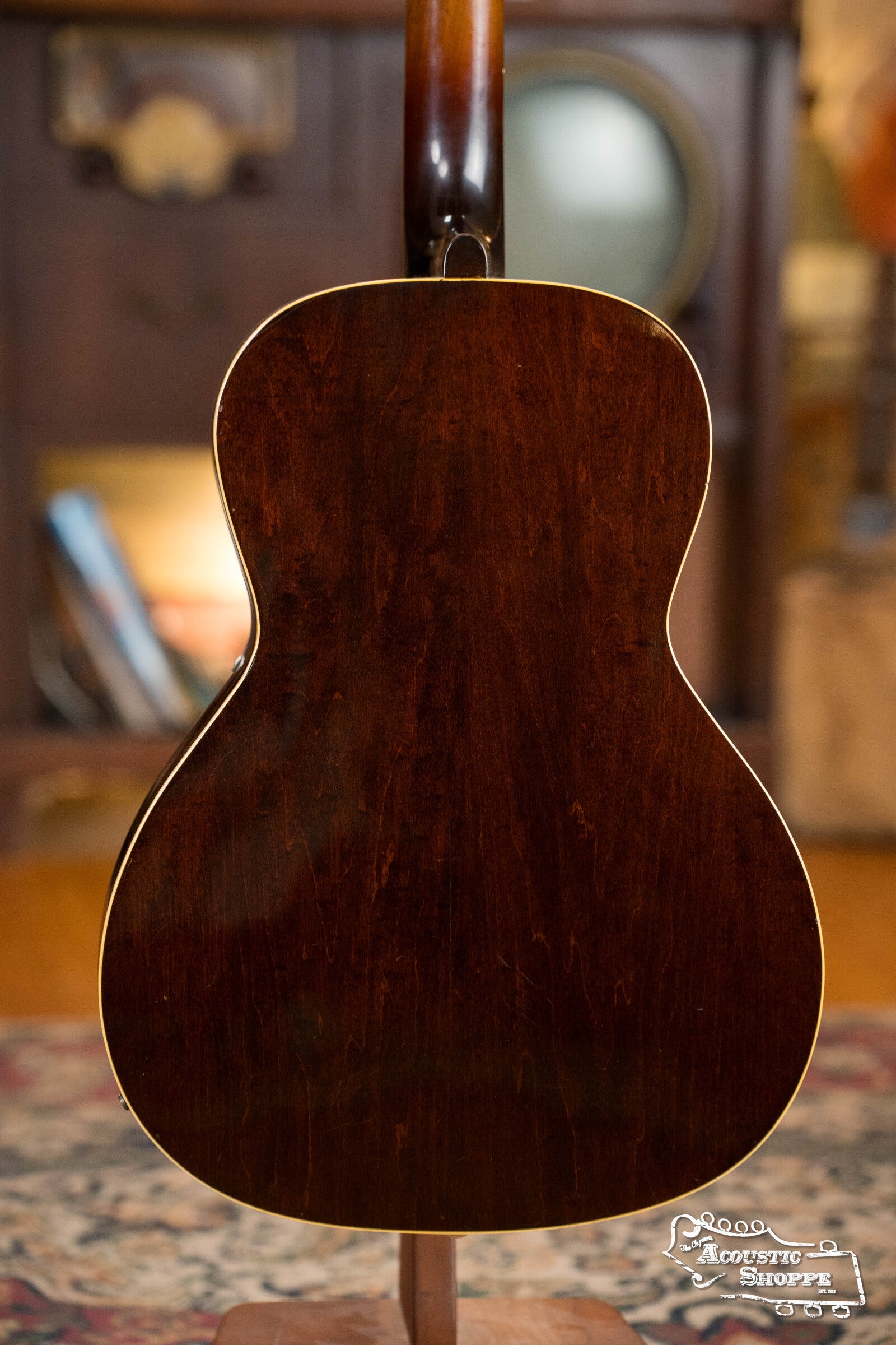A (Used) 1942 Gibson ES-125 by Gibson stands upright on a stand, showcasing its rich dark wood finish. The softly blurred background features a wooden cabinet and books.